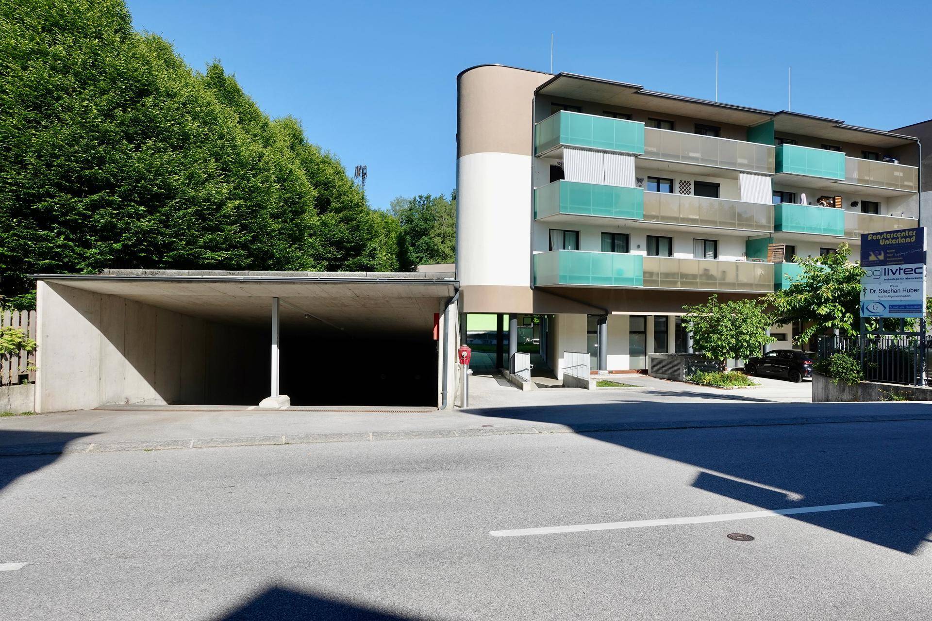 Mehrstöckiges Gebäude mit Balkonen, grünen Glasgeländern und einer Einfahrt zur Tiefgarage.