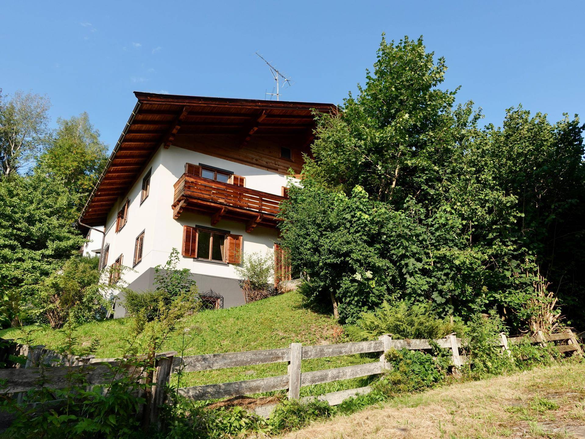 Haus auf einem Hügel mit Holzbalkonen und dichter Vegetation in ländlicher Umgebung.