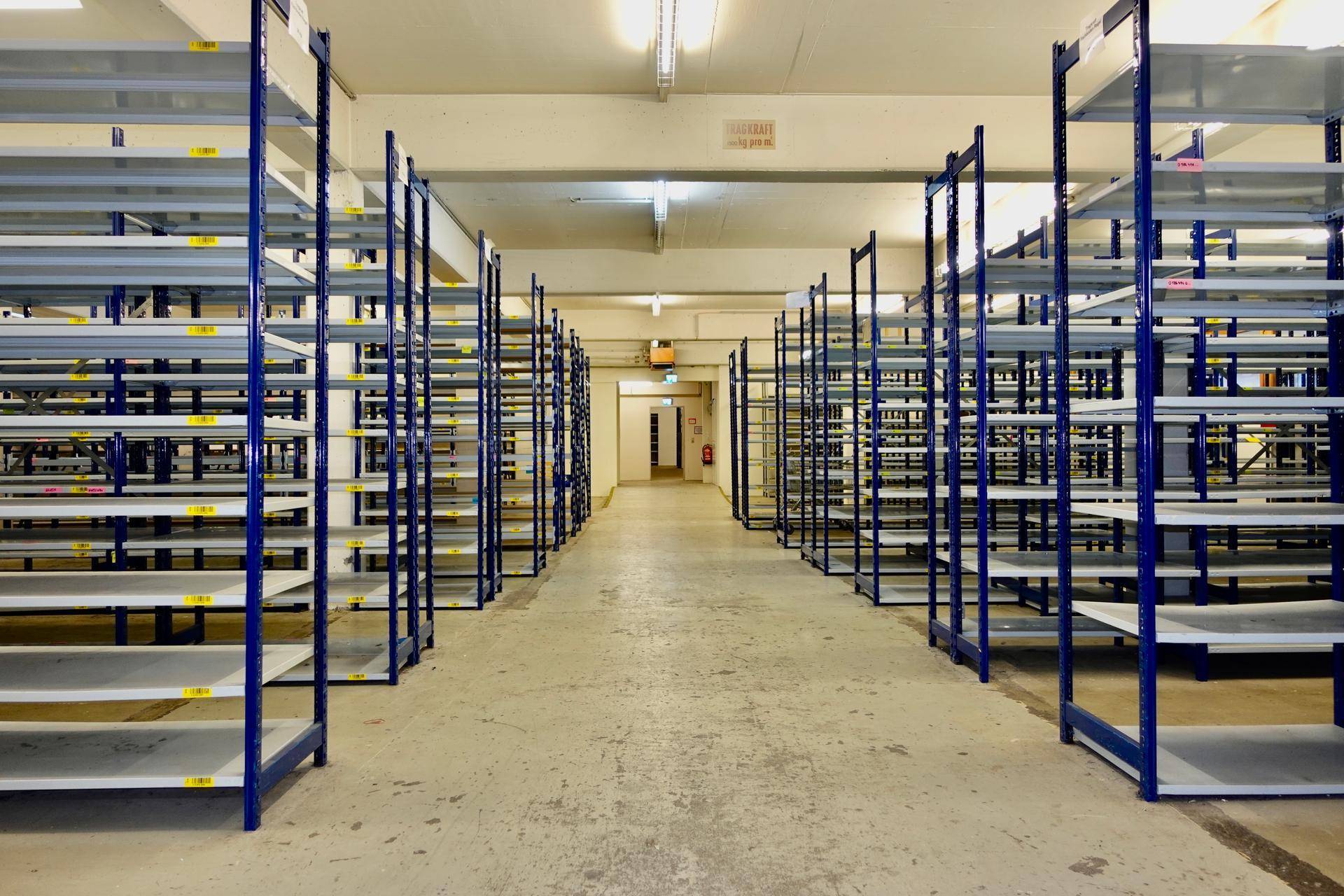 Langer Gang in einer Lagerhalle mit vielen leeren blauen Metallregalen und Betonboden.