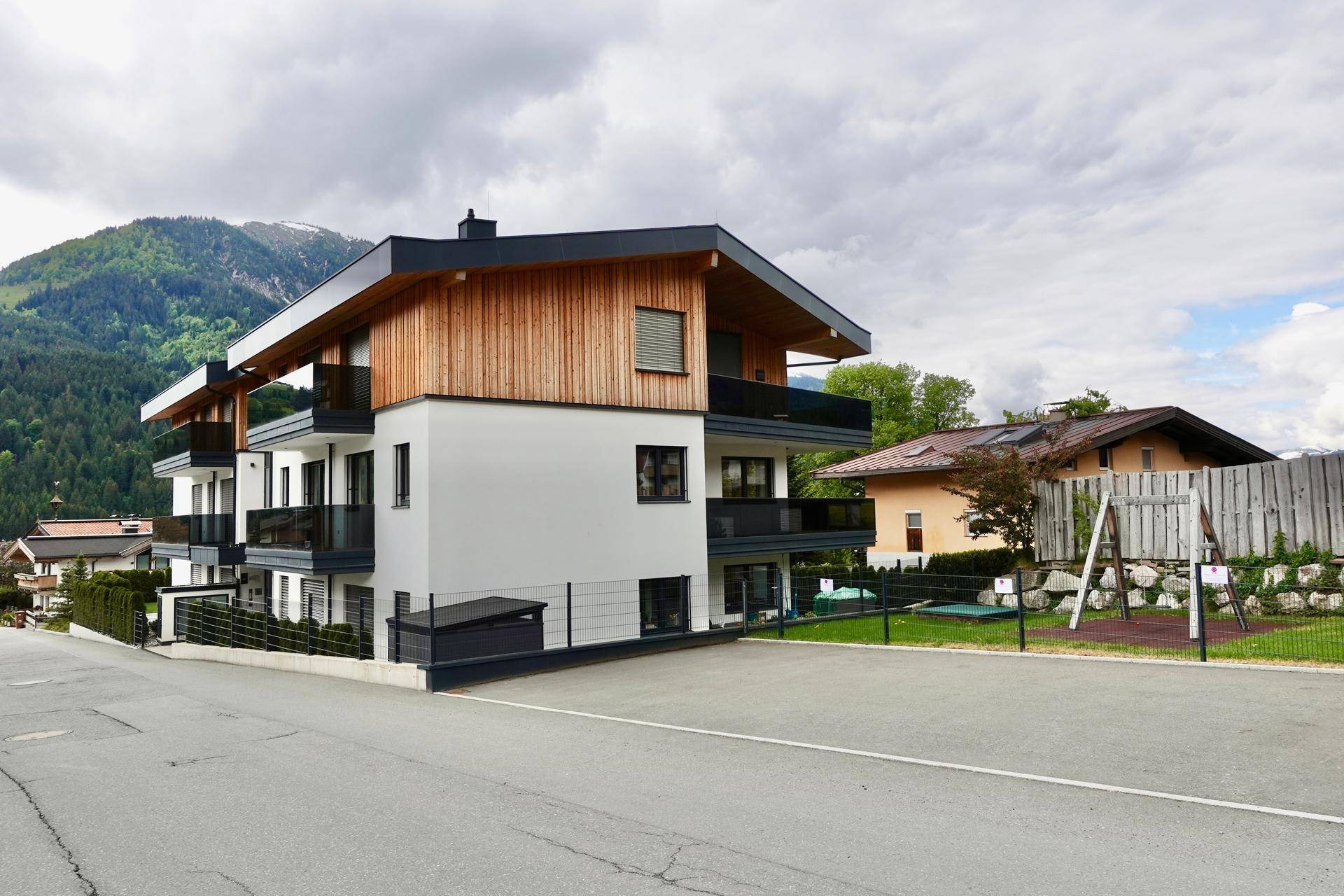 Neues Mehrfamilienhaus mit modernen Balkonen und einem Spielplatz im Grünen, umgeben von Bergen.