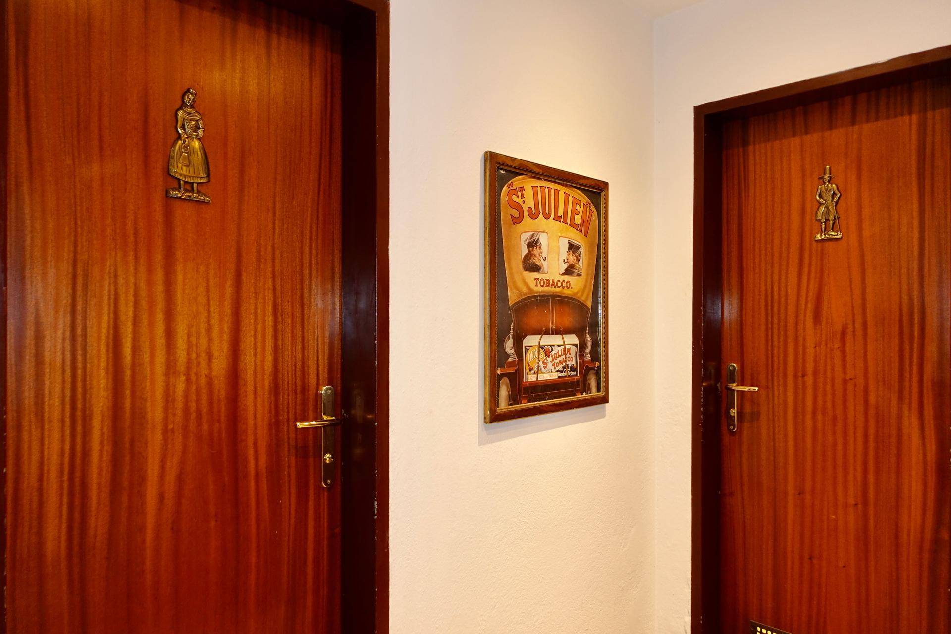 Zwei Holztüren mit traditionellen Beschlägen und einem Vintage-Werbeplakat an der Wand, die zu den Toiletten führen.