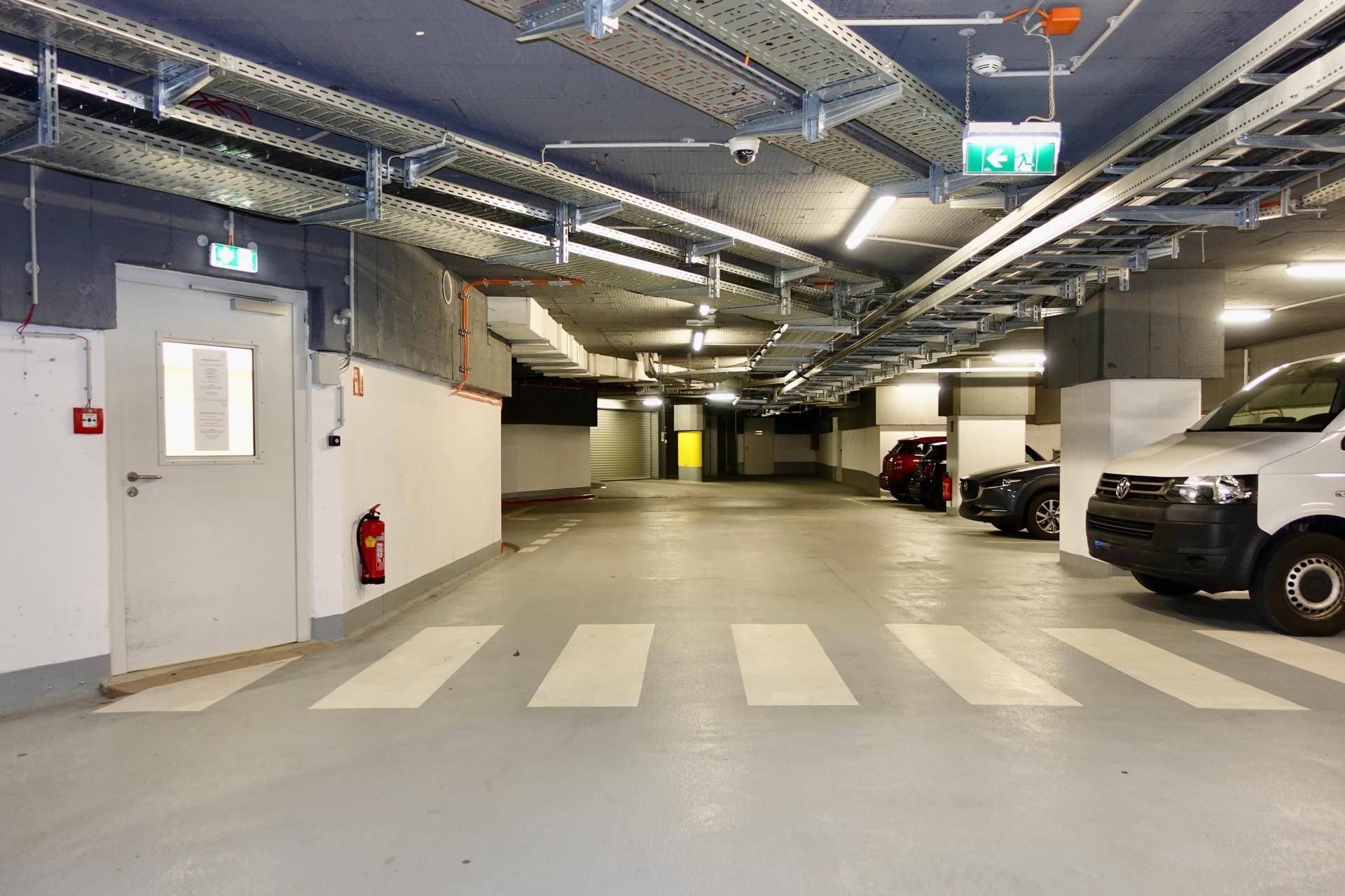 Geräumige Tiefgarage mit mehreren Fahrzeugen, Sicherheitsausrüstung und Fluchtwegbeschilderung an den Wänden.