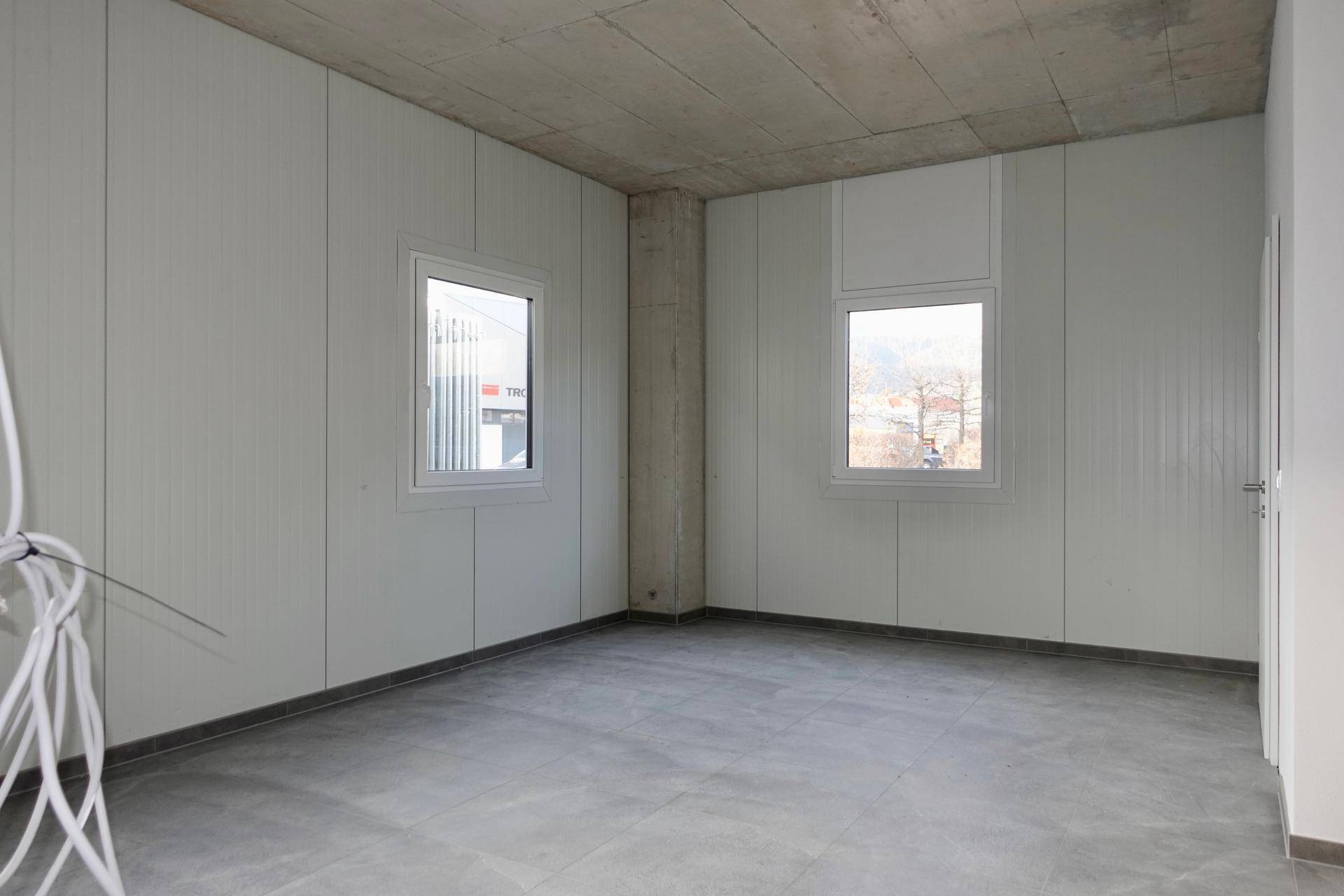 Leerer Raum mit zwei Fenstern, grauen Fliesen und einer unverputzten Betondecke.