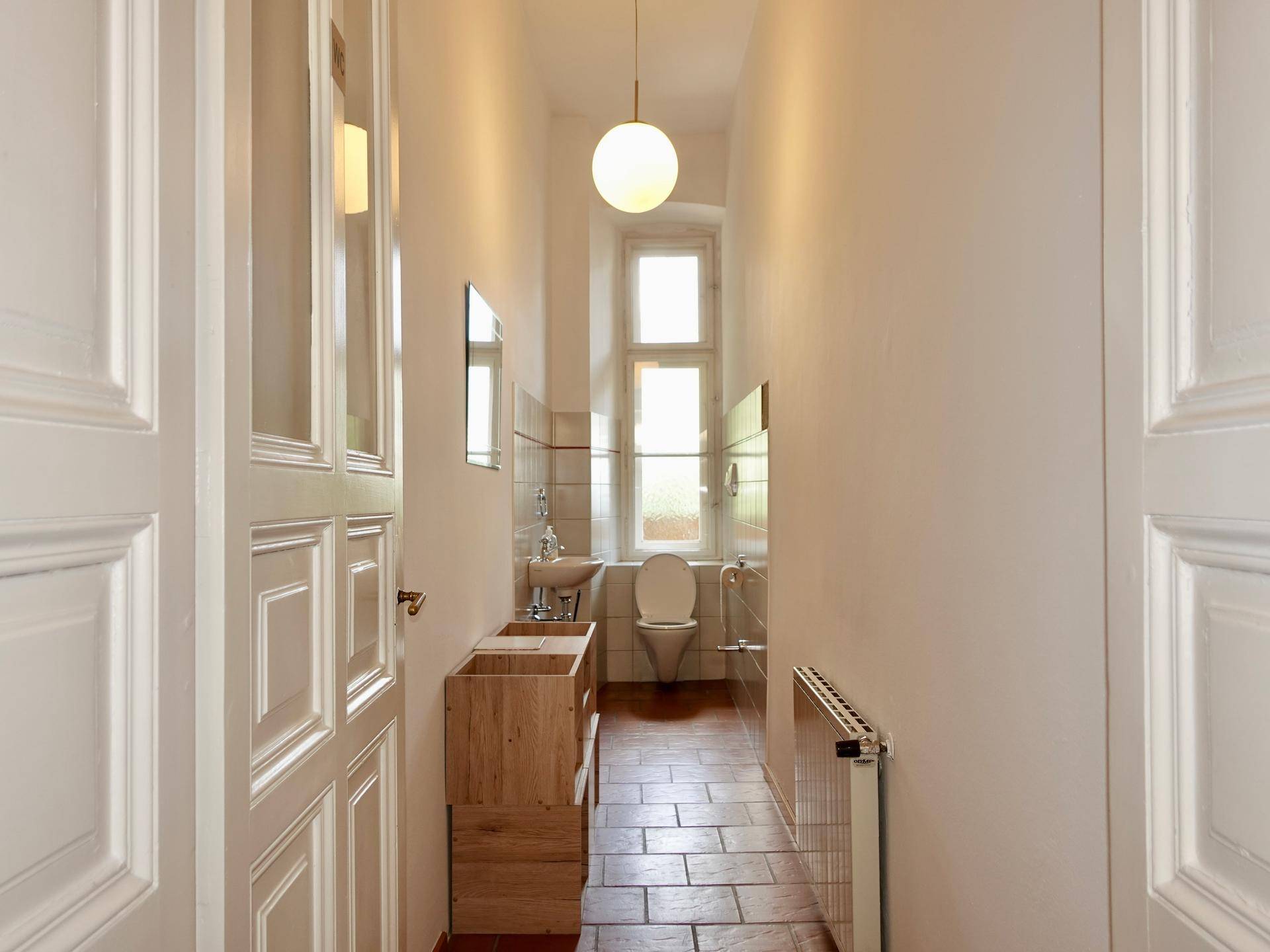 Schmale Gästetoilette mit Fenster, Waschbecken, WC und Terrakottafliesen, gut beleuchtet.