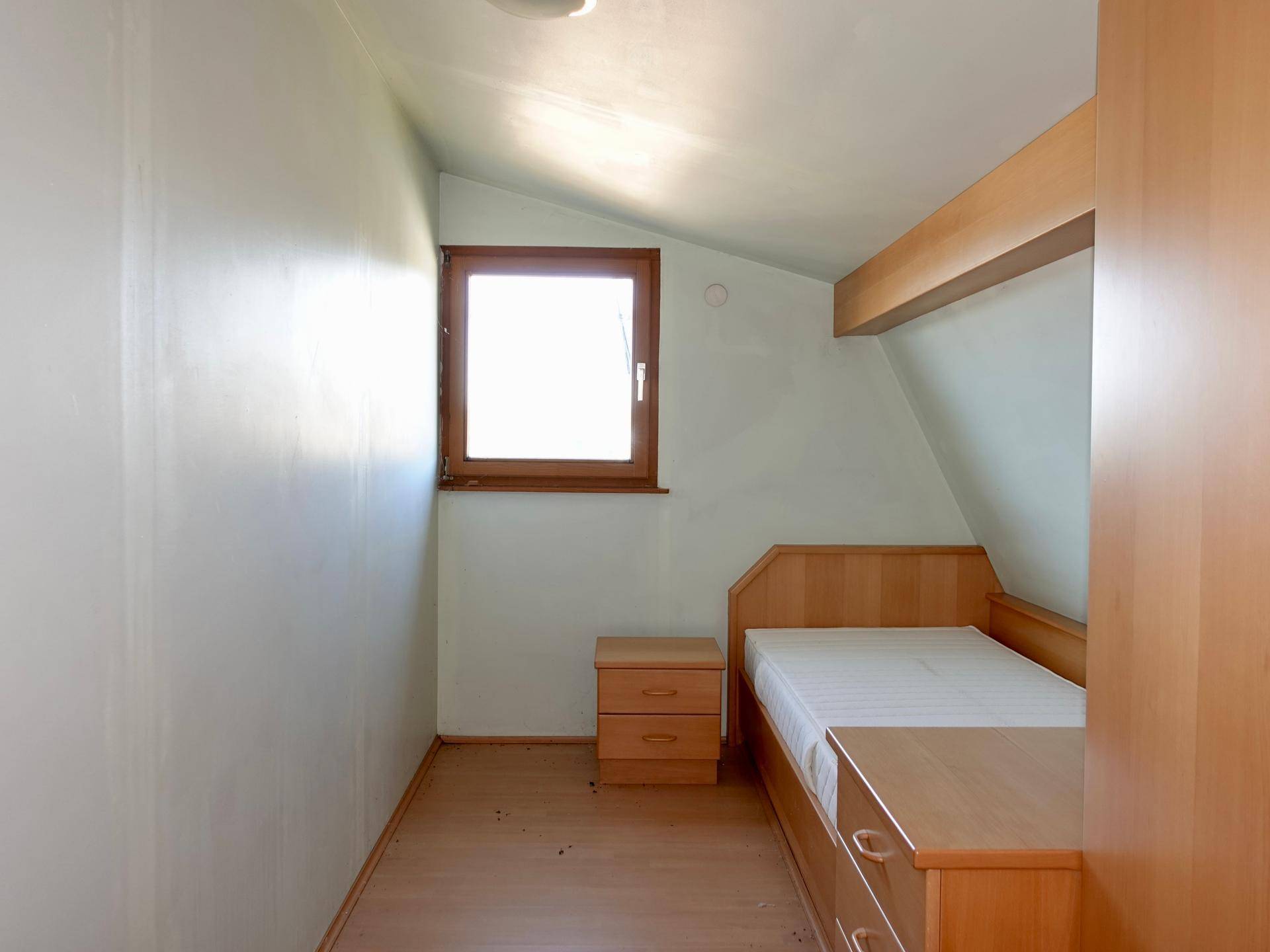 Kleines Schlafzimmer mit Einzelbett, Holzmöbeln und Dachschräge, hell beleuchtet.