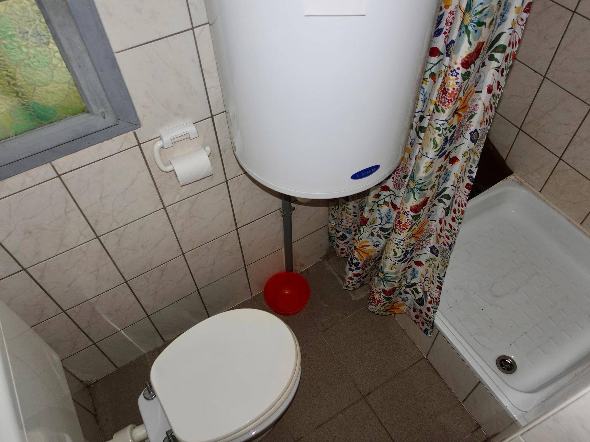 Kleines Badezimmer mit WC, Dusche und Warmwasserboiler, gefliest.