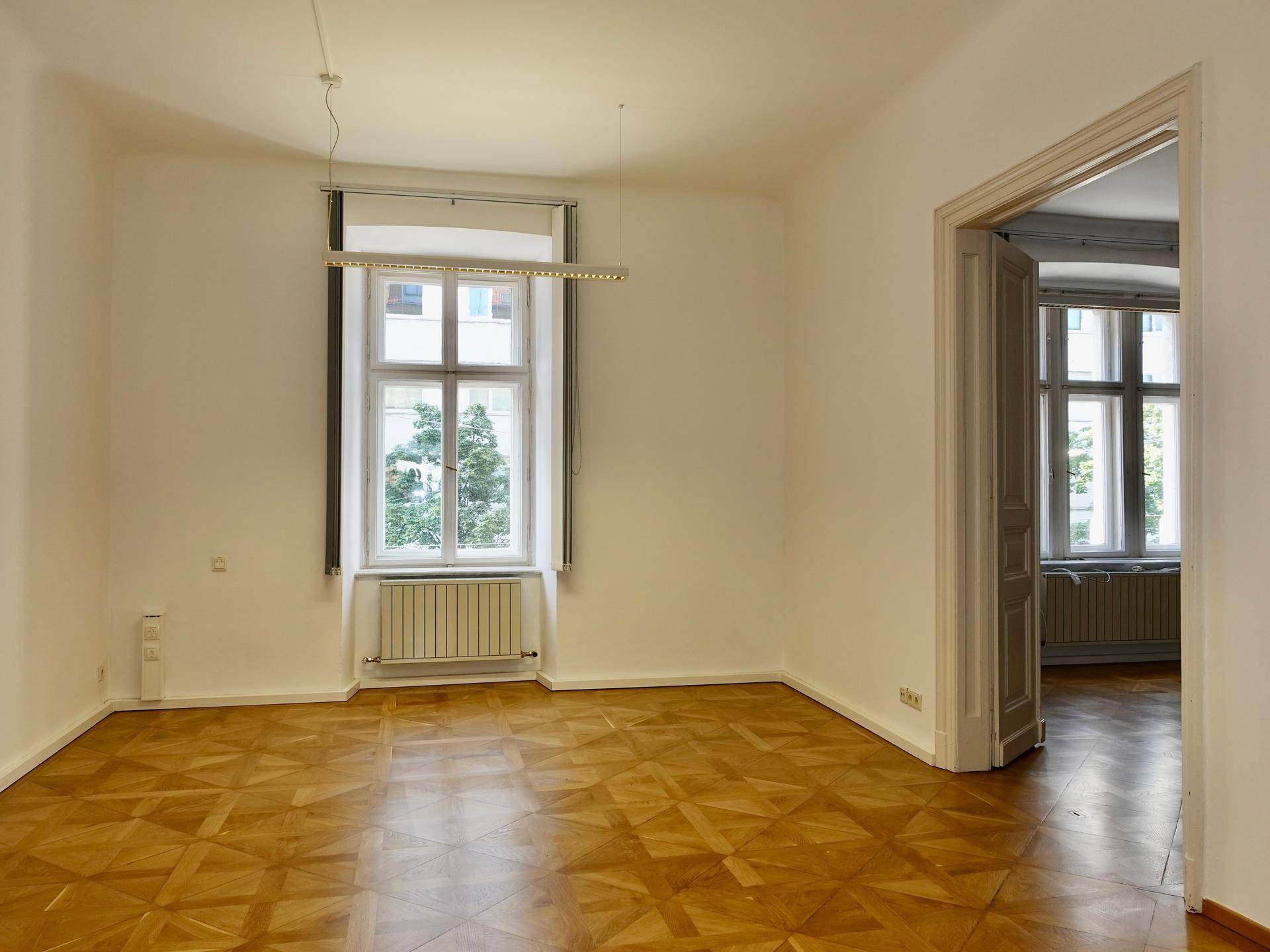 Heller Raum mit Fischgrätparkett, großem Fenster und Blick in einen weiteren Raum mit ähnlichem Stil.