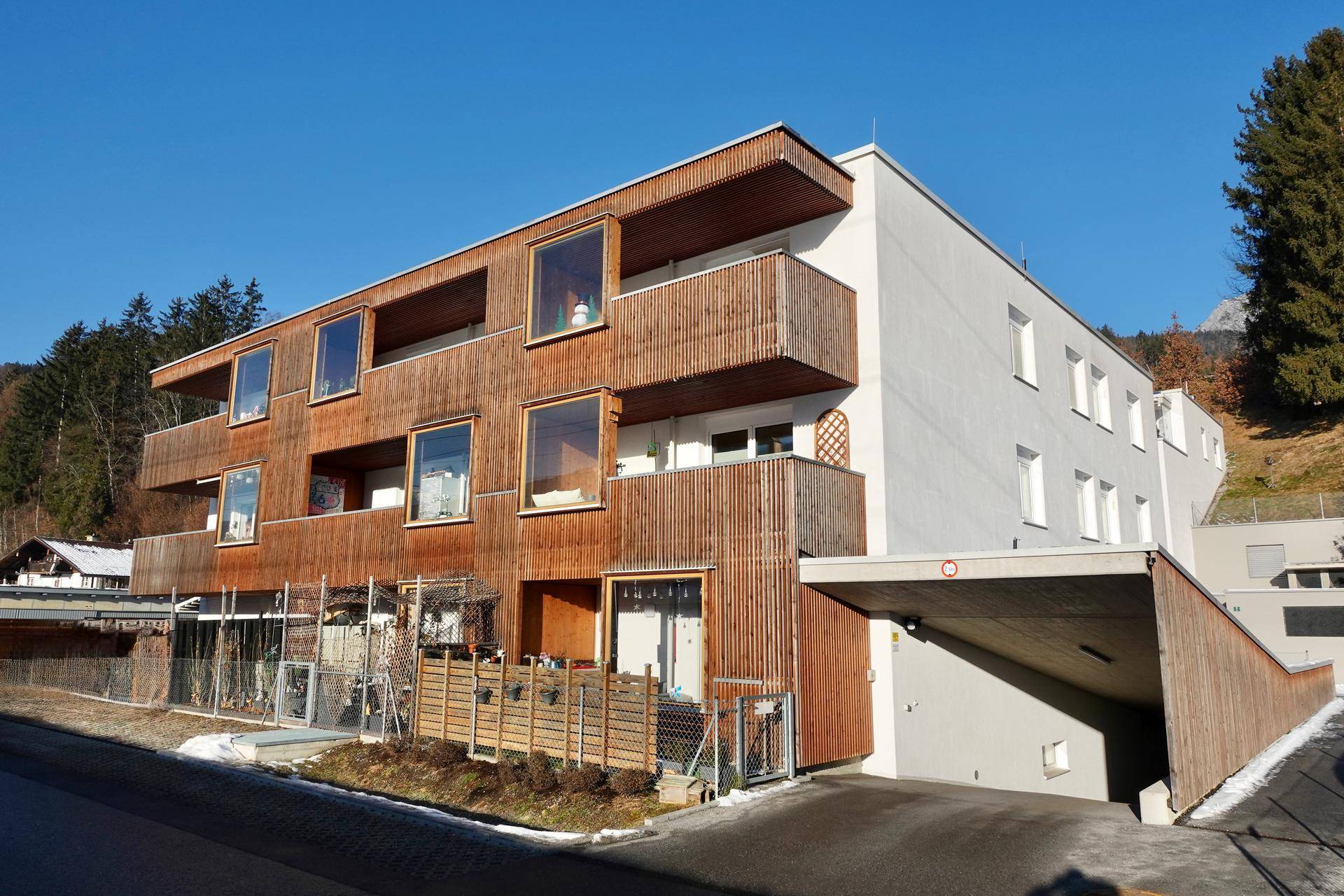 Modernes Mehrfamilienhaus mit Holzfassade, Balkonen und Tiefgaragenzufahrt vor bergiger Kulisse.