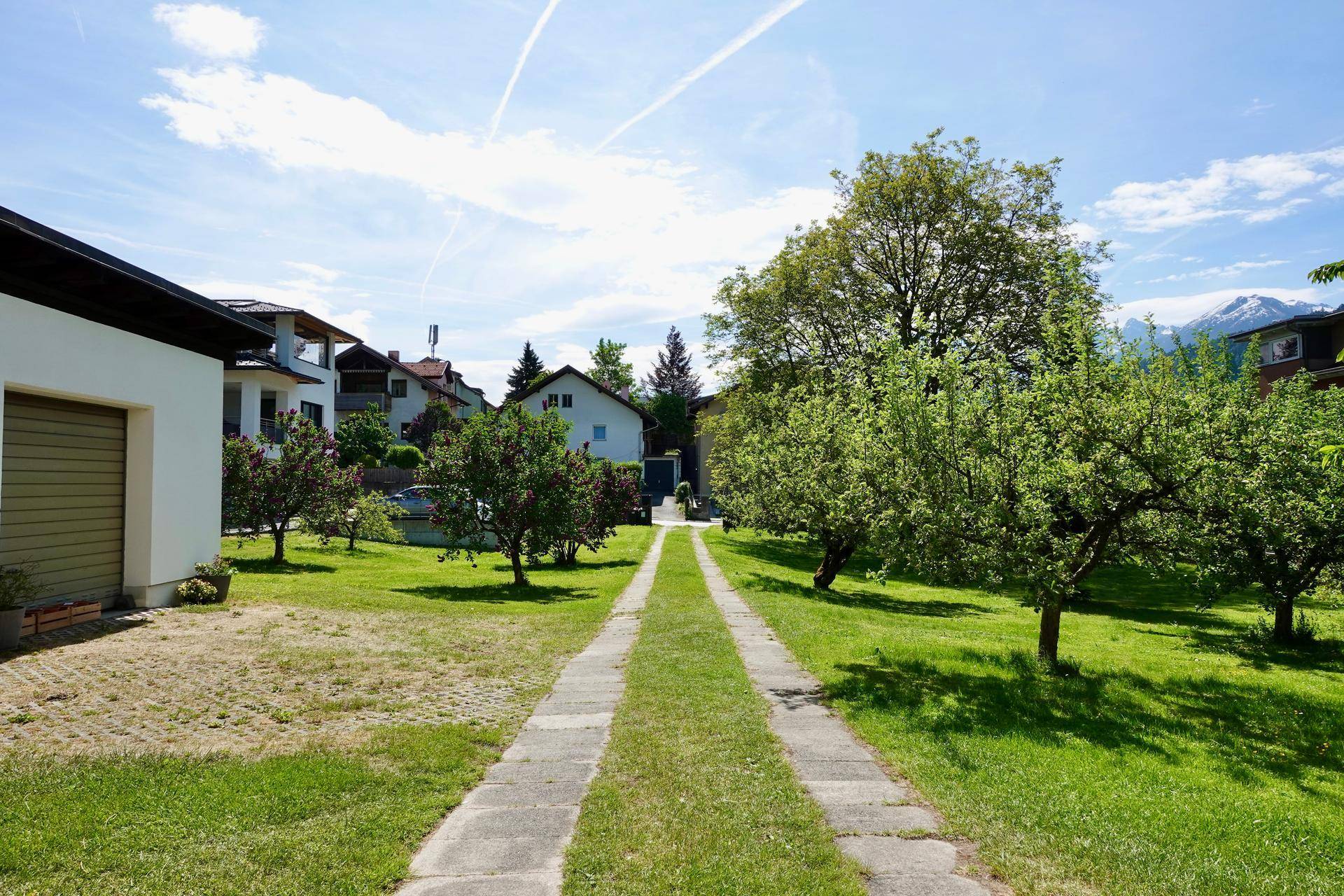 Langer gepflasterter Weg durch einen grünen Garten mit Obstbäumen und Blick auf die Berge.