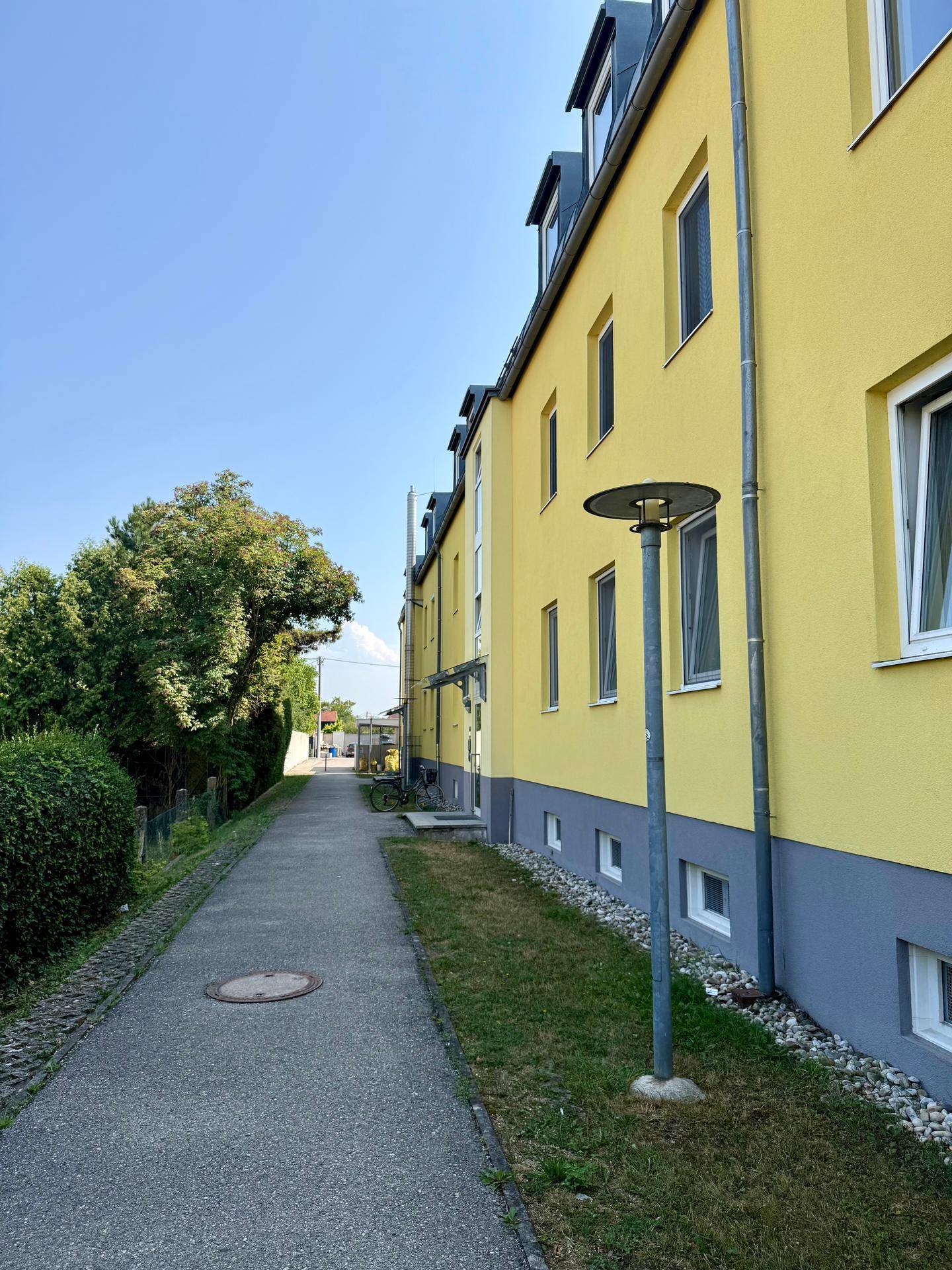 Seitenansicht eines gelben Mehrfamilienhauses mit Gehwegen und grüner Bepflanzung.