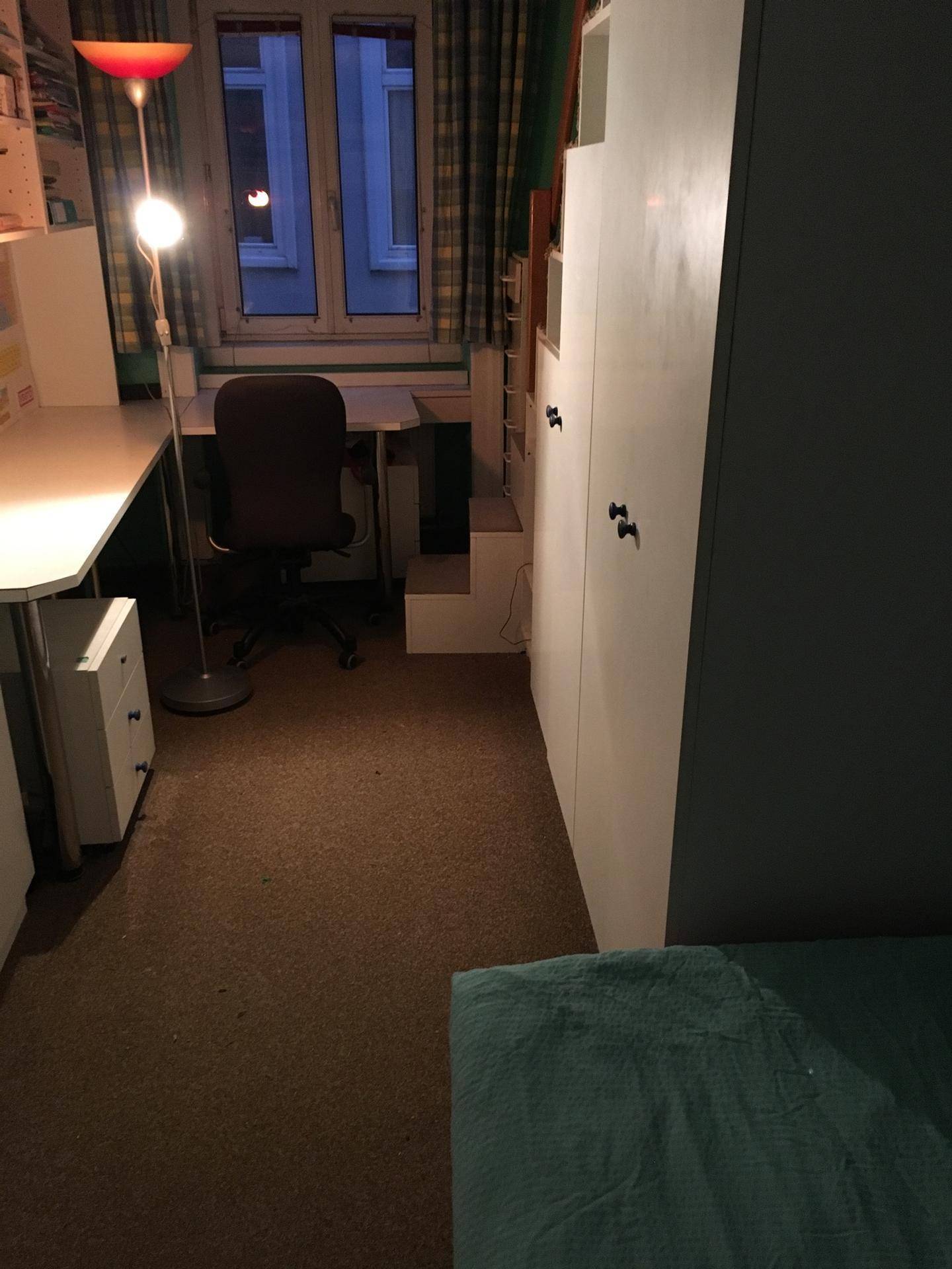Multifunktionaler Raum mit Schreibtisch, Stuhl, Stehlampe und einem Fenster, das Tageslicht hereinlässt.