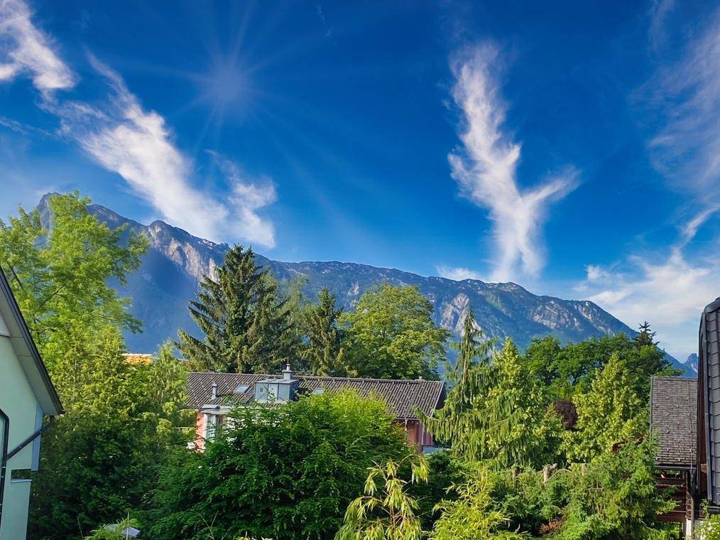 Malerischer Bergblick über grüne Baumwipfel und Dächer unter strahlend blauem Himmel.