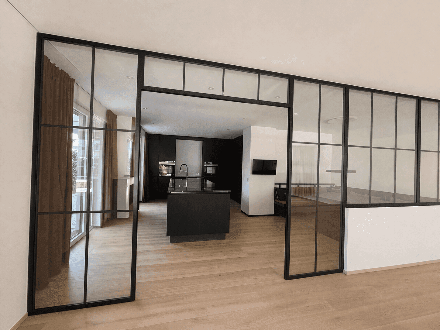 Moderner offener Wohnbereich mit Holzboden und eleganten schwarzen Glastrennwänden zur Küche.