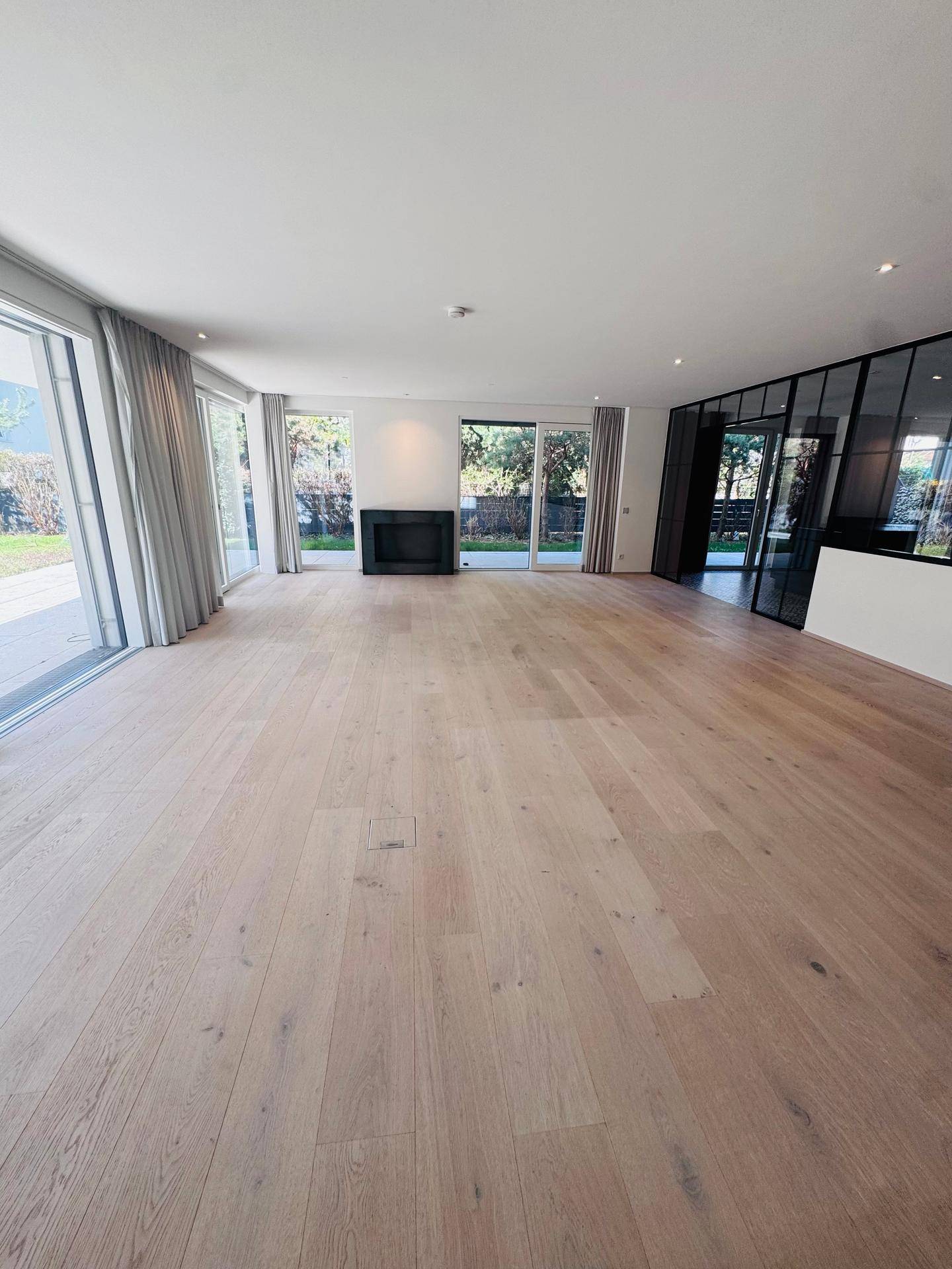 Geräumiges Wohnzimmer mit hellem Holzboden, modernem Kamin und großen Glasfronten zum Garten.