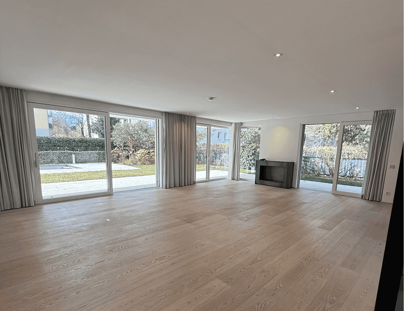 Heller, leerer Wohnraum mit Holzboden, Kamin und raumhohen Fenstern, die viel Licht hereinlassen.