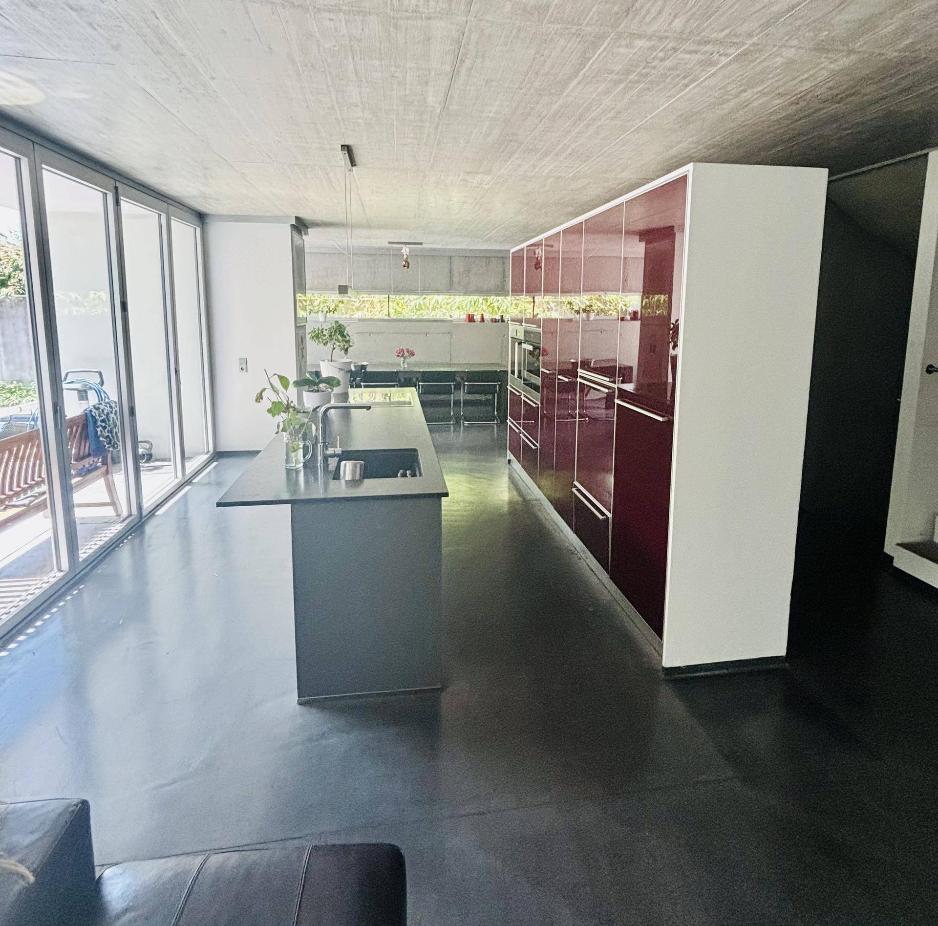 Moderne Küche mit roten Hochglanzschränken, Kücheninsel und großflächigen Fenstern zum Außenbereich.