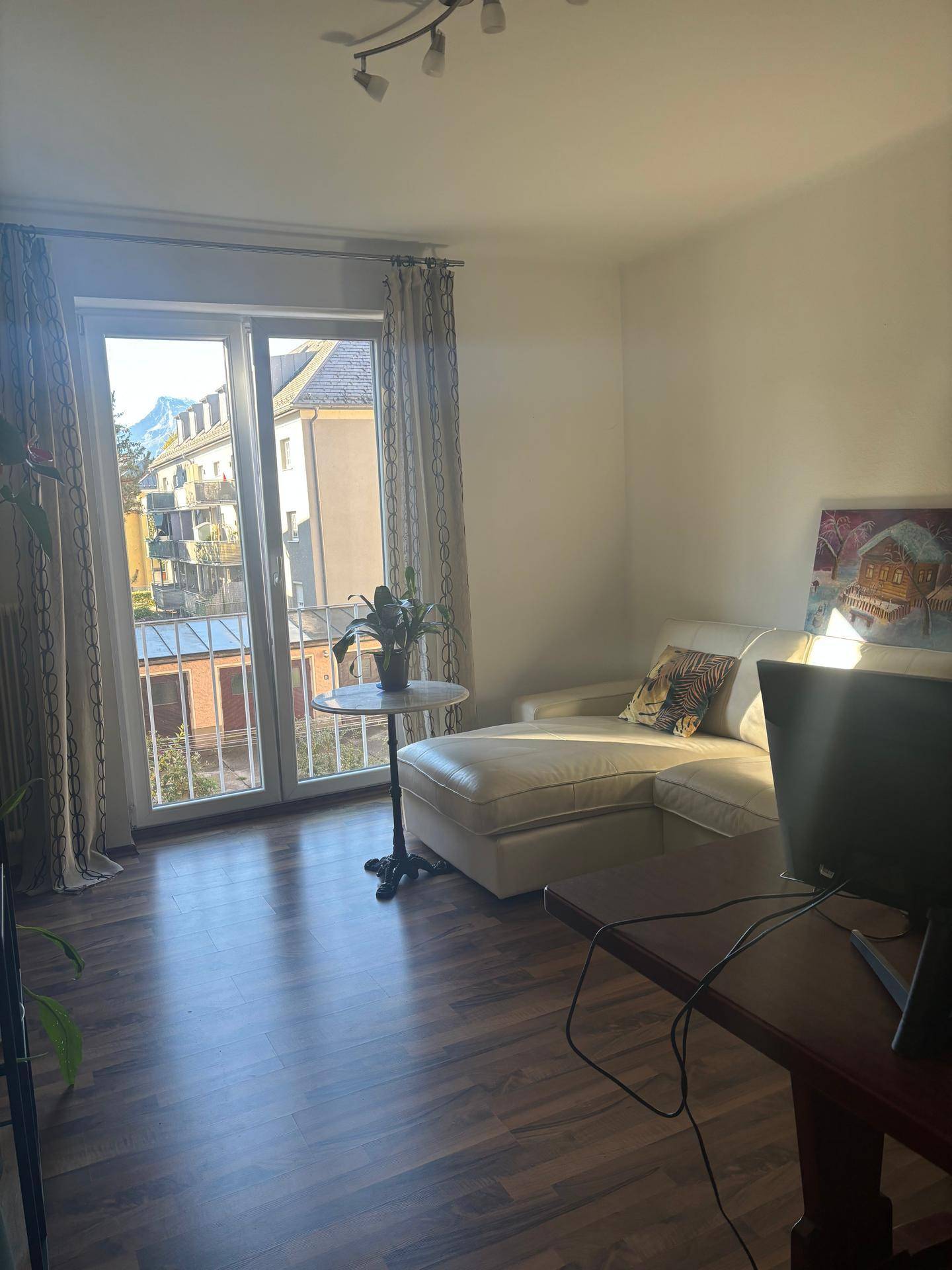 Helles Wohnzimmer mit beiger Chaiselongue, dunklem Laminatboden und Zugang zu einem kleinen Balkon mit Blick auf die Umgebung.