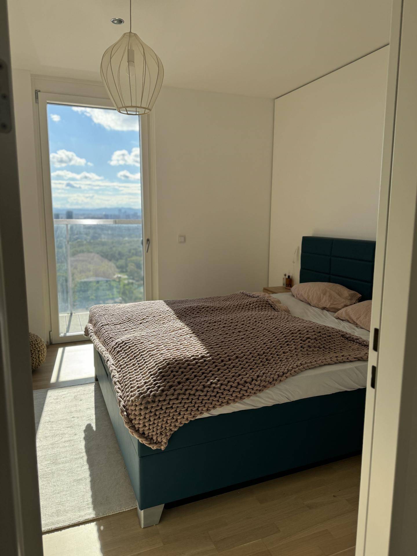 Sonniges Schlafzimmer mit großem Fenster, Holzboden und weitem Blick auf die Stadt.