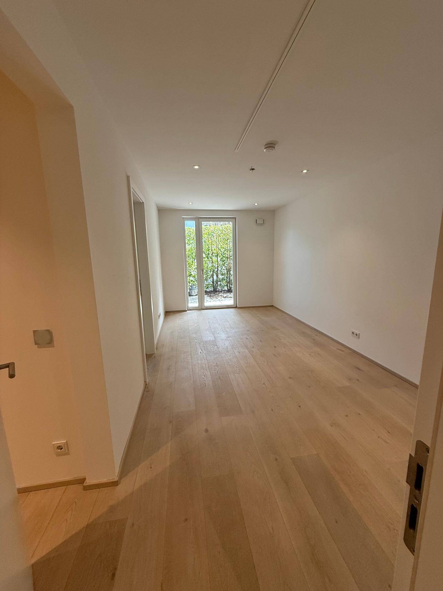 Helles, leeres Schlafzimmer mit Holzboden und großer Glastür, die viel Tageslicht hereinlässt.