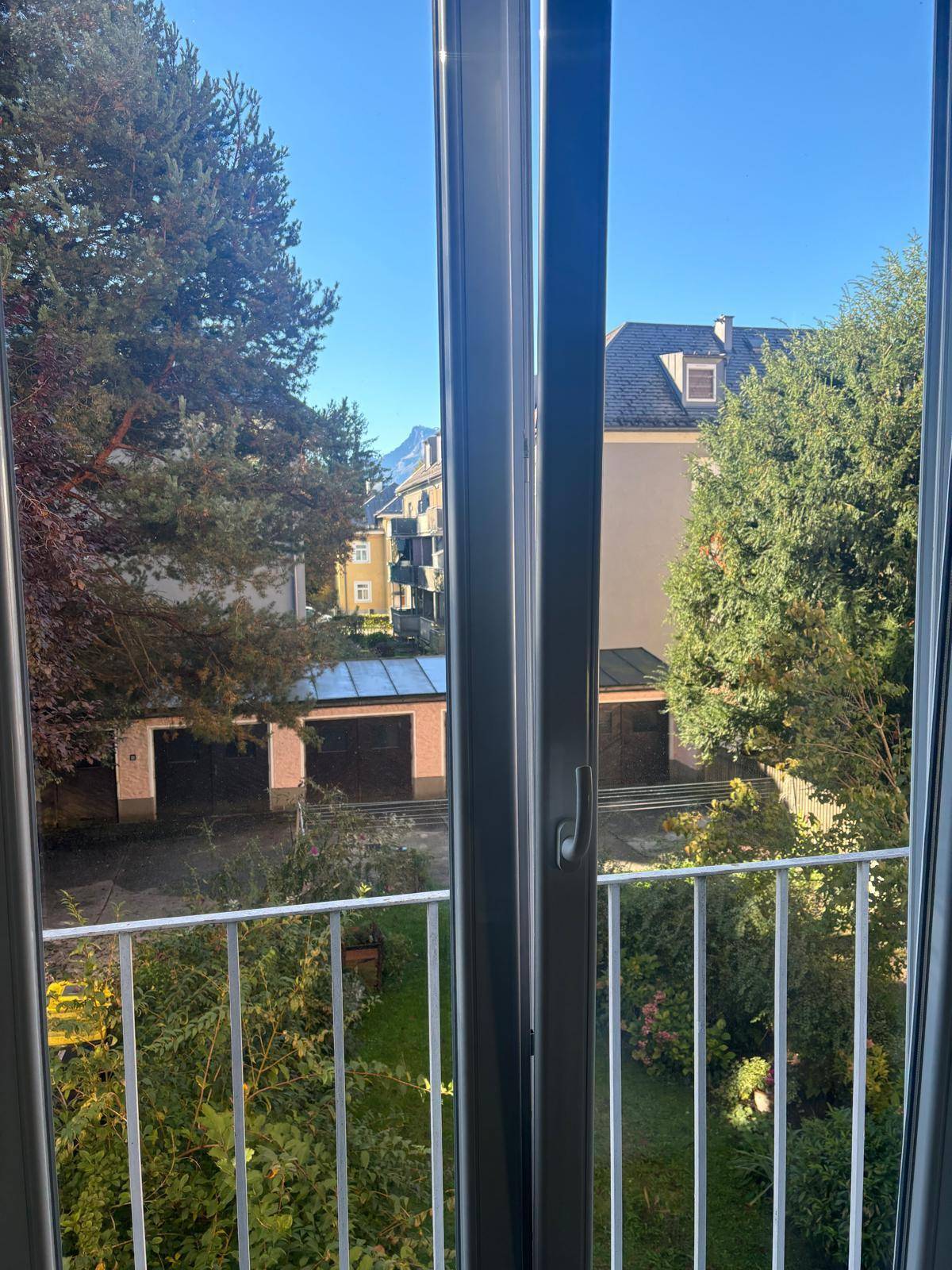 Blick aus dem Fenster auf einen begrünten Innenhof mit Garagen und umliegenden Gebäuden, im Hintergrund Berge sichtbar.