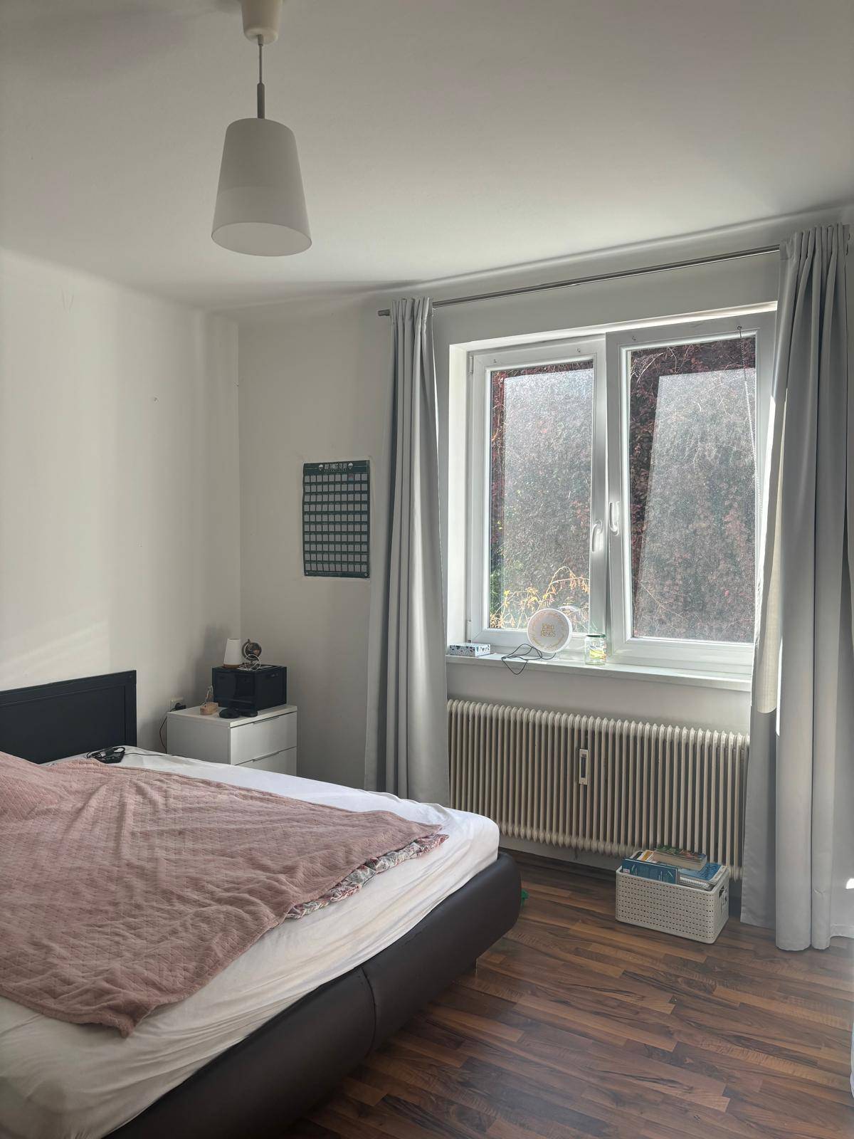 Schlafzimmer mit dunklem Laminatboden, großem Fenster und Heizkörper unter der Fensterbank.