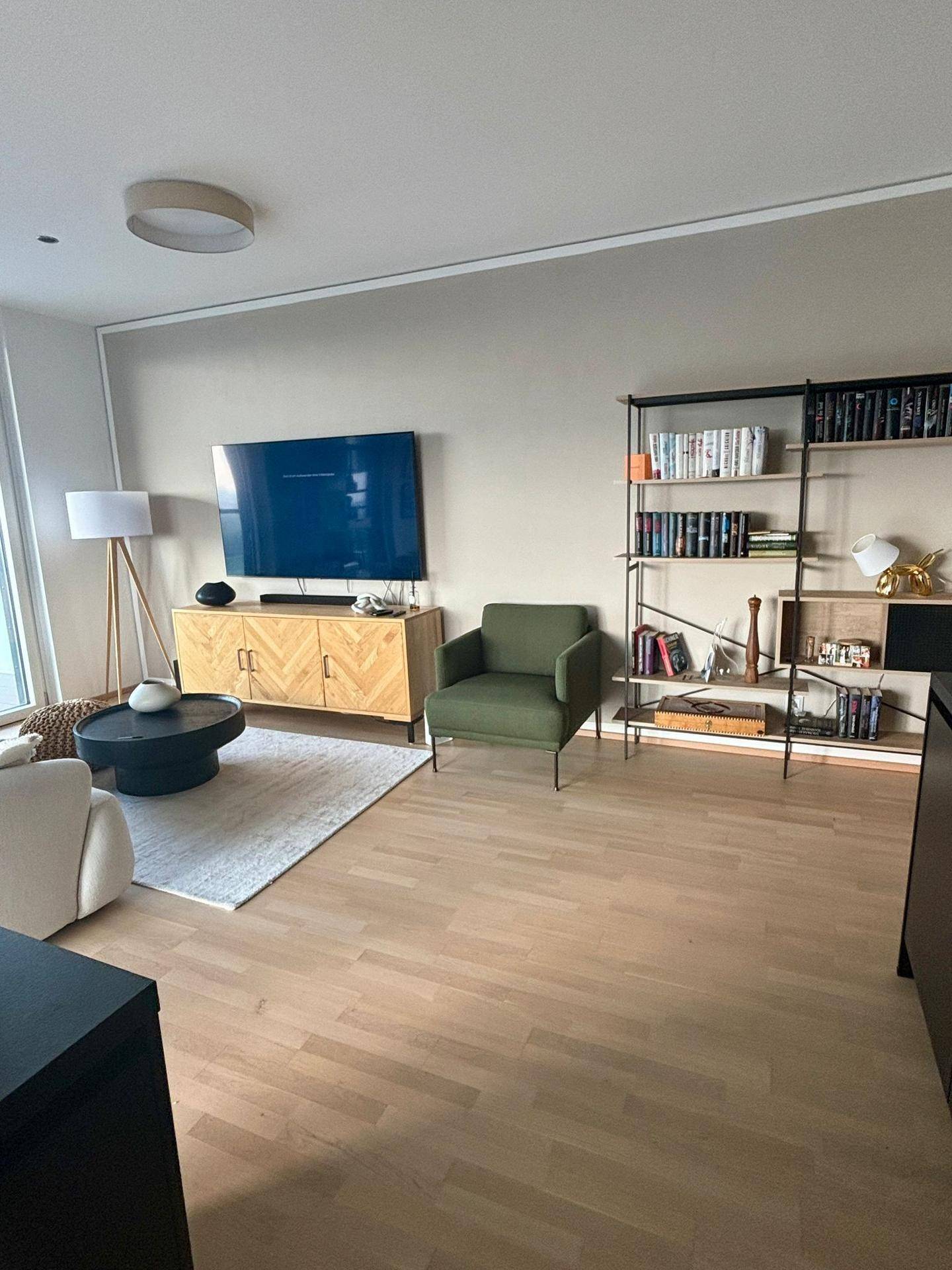 Modernes Wohnzimmer mit Fernseher, grünem Sessel, Bücherregal und Holzboden.