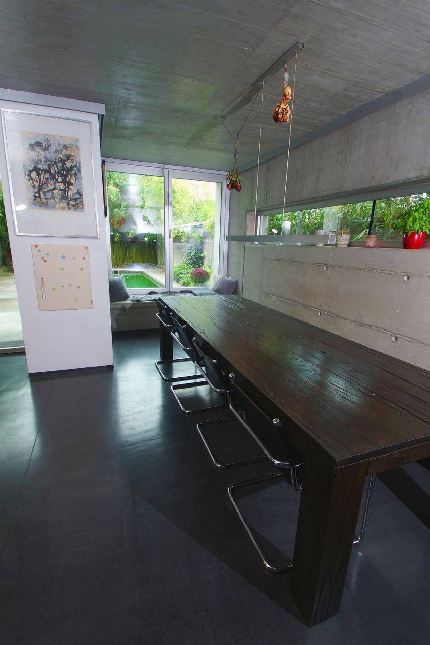 Essbereich mit langem Holztisch, Betonwänden und großen Fenstern mit Blick auf den Garten.