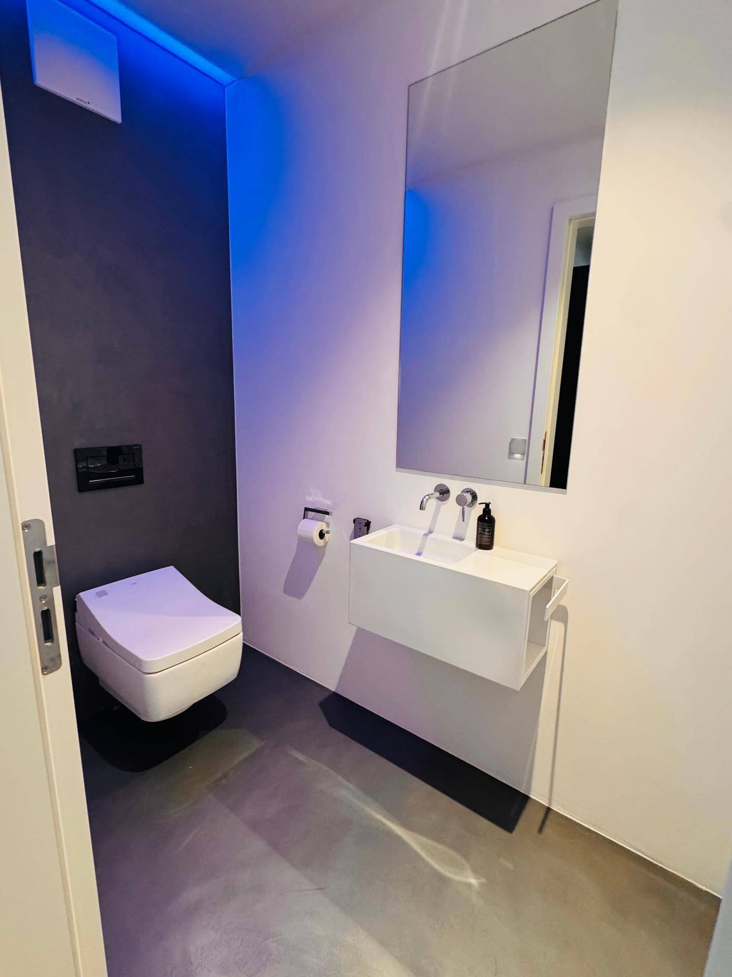 Modernes Gäste-WC mit Wand-WC, kleinem Waschbecken und indirekter blauer Beleuchtung.