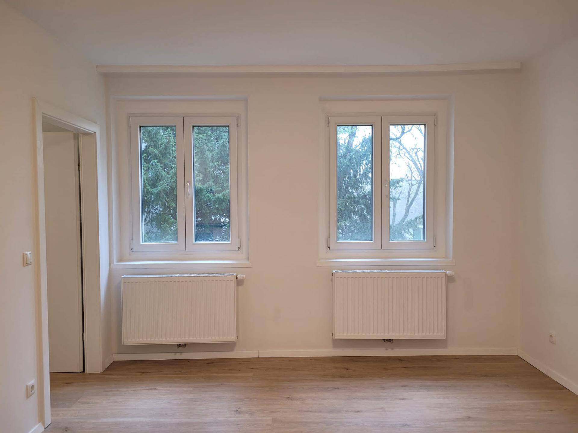Heller Raum mit zwei Fenstern, Laminatboden und Ausblick auf Bäume.