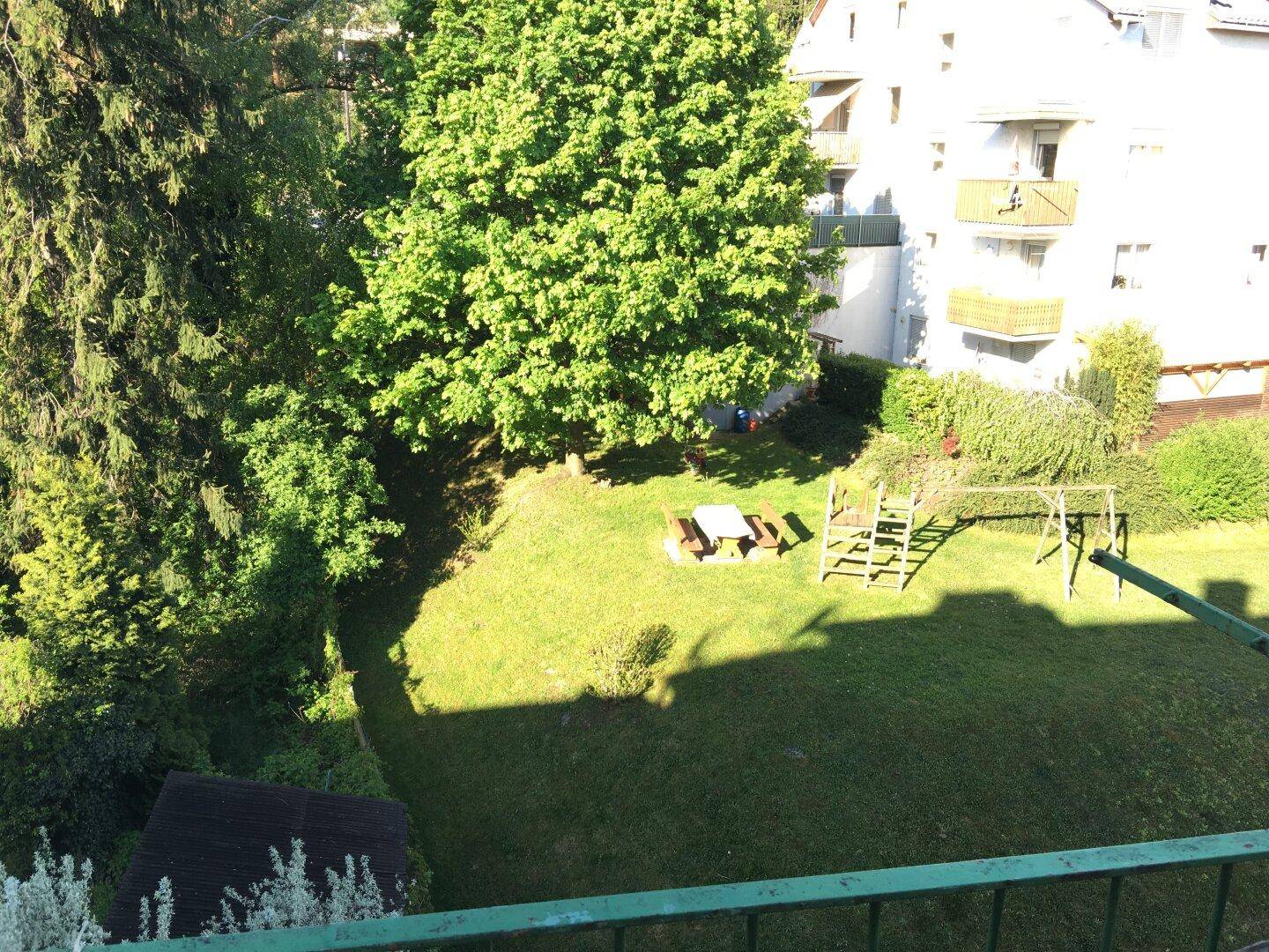 Sonniger Blick auf einen grünen Gemeinschaftsgarten mit hohen Bäumen, Picknicktisch und Kinderspielgeräten.