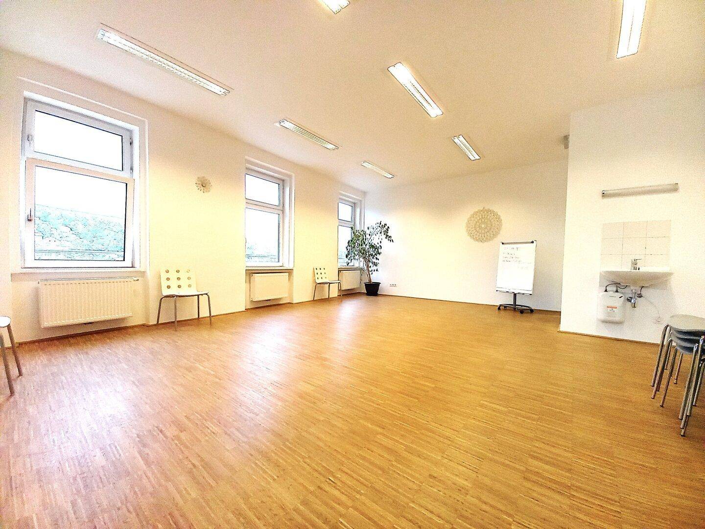 Großer, heller Raum mit Holzfußboden, mehreren Fenstern und einer kleinen Küchenzeile, ideal für Bürozwecke.