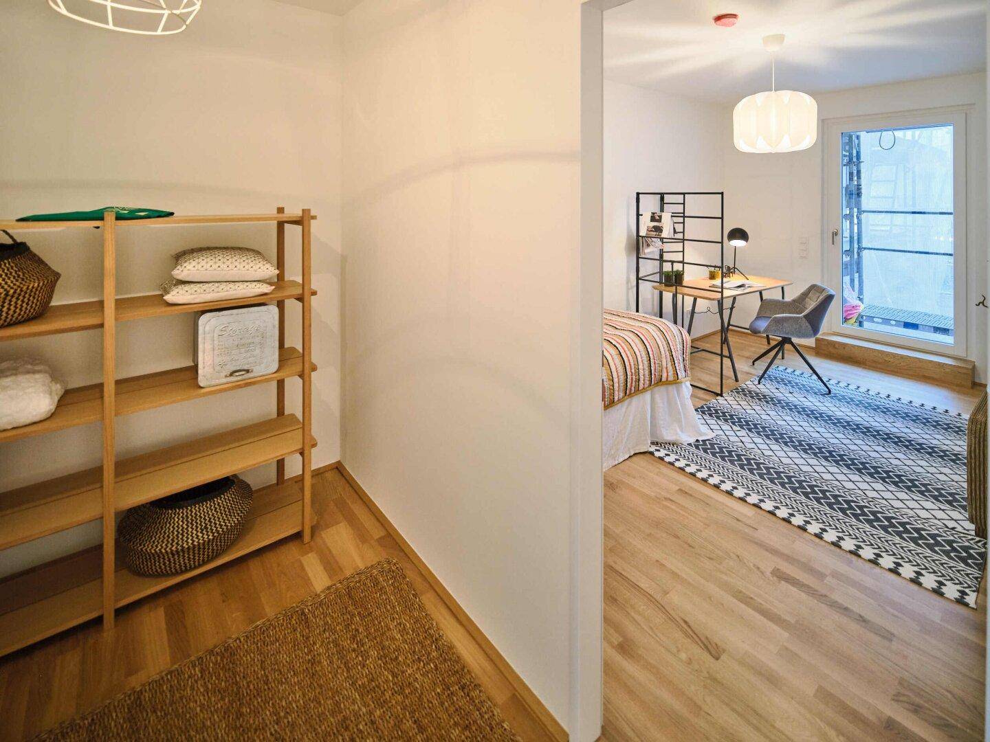 Heller Flur mit Holzregal und Blick in ein angrenzendes Zimmer mit Holzboden.