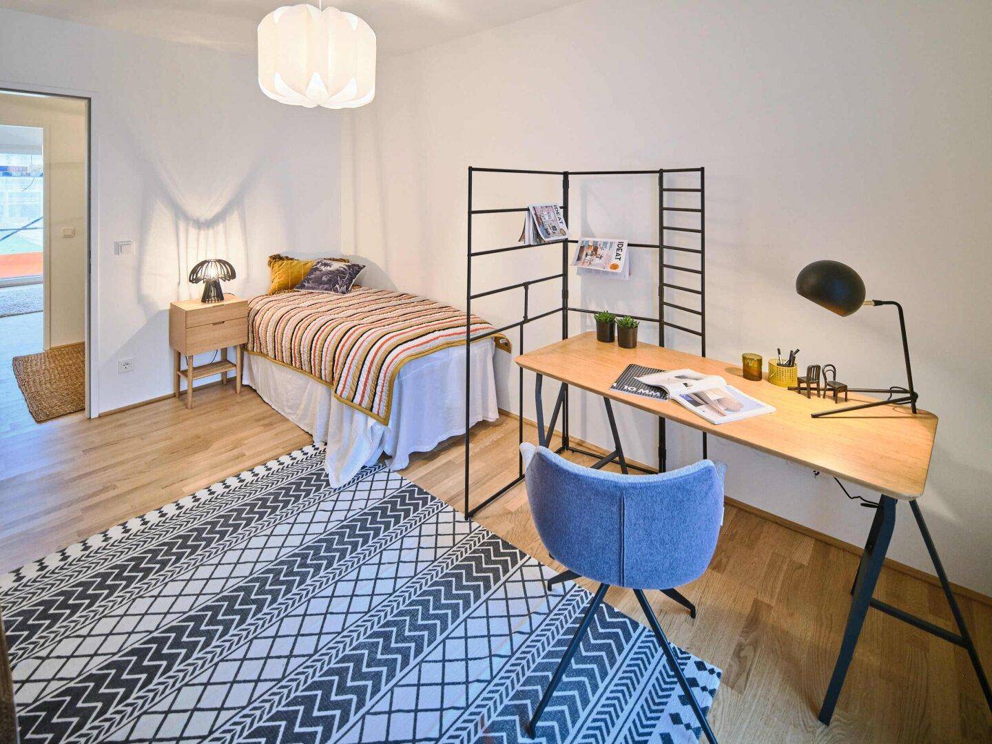 Helles Schlafzimmer mit Schreibtisch, Stuhl und Einzelbett mit farbiger Bettdecke.