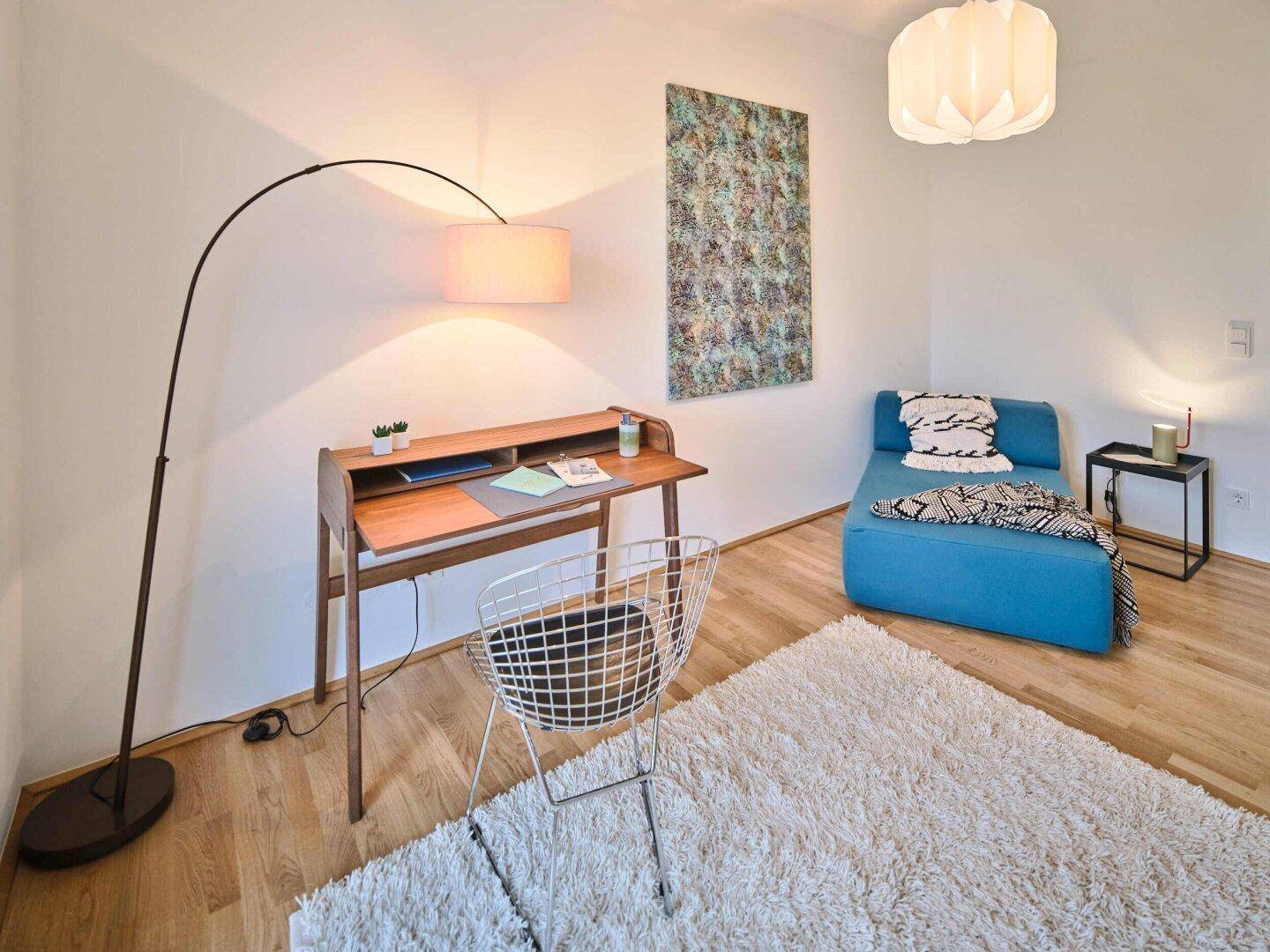 Arbeitszimmer mit Schreibtisch, Stehlampe und kleinem blauen Sofa.