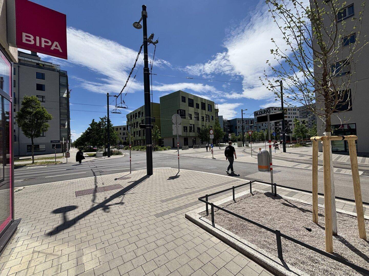 Belebte Straßenkreuzung in einem modernen Stadtviertel mit Fußgängern und umliegenden Gebäuden.