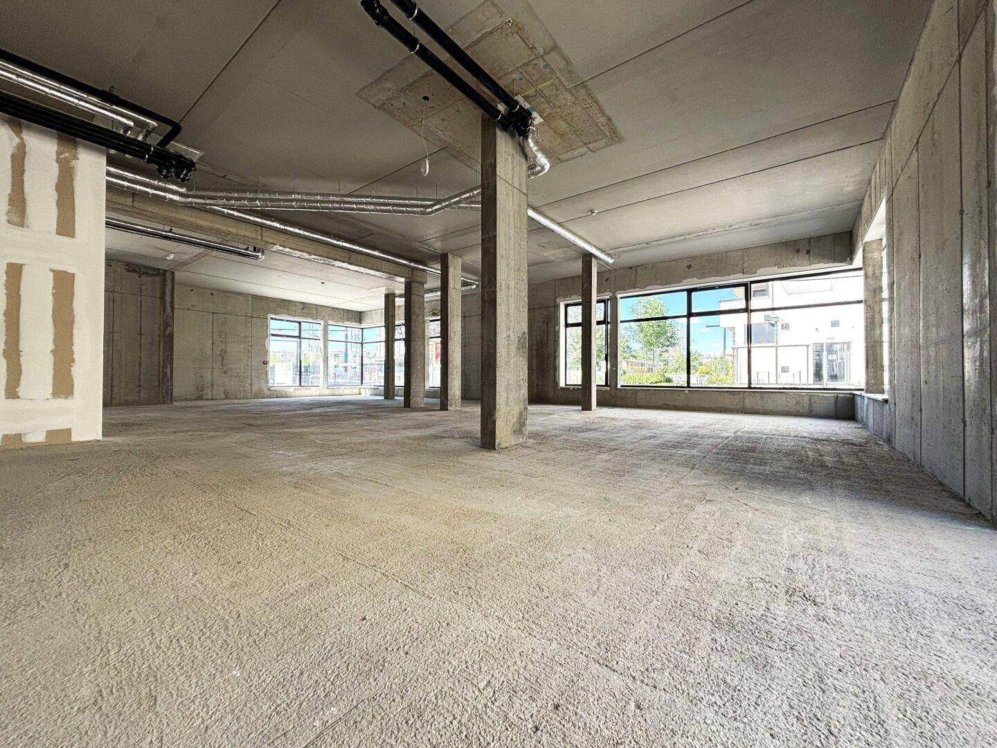Geräumiger, unfertiger Innenraum mit Betonwänden und -boden, großen Fenstern und sichtbaren Installationen.