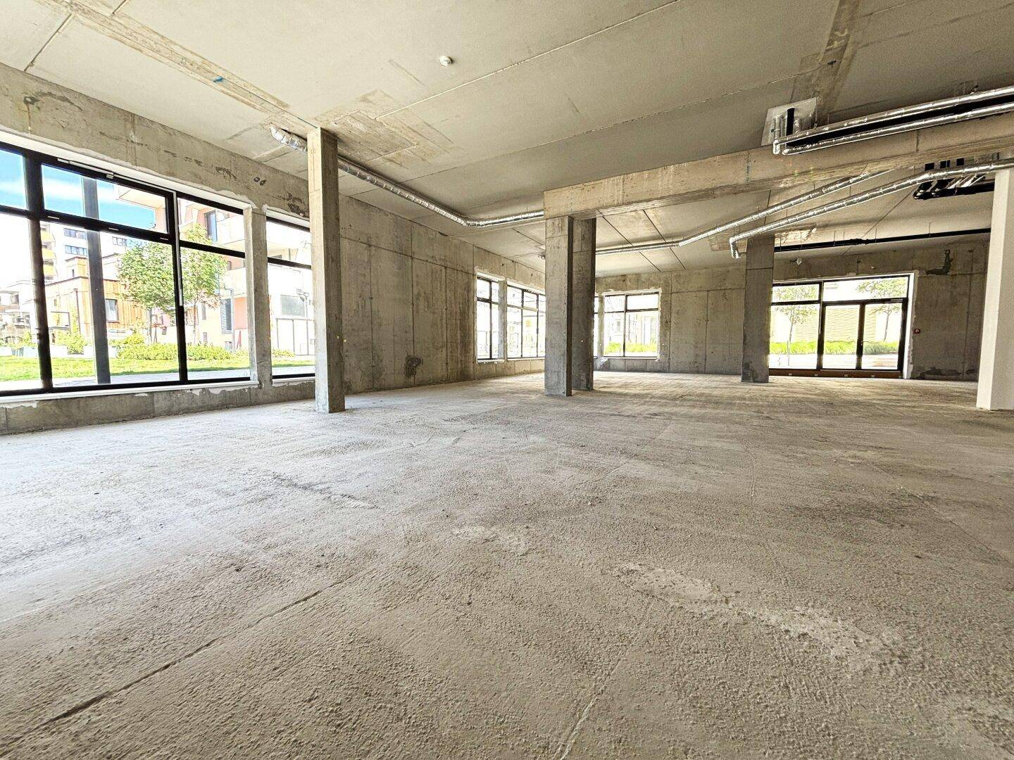 Weitläufiger, unfertiger Innenraum mit Betonboden und -wänden, großen Fenstern und viel Tageslicht.