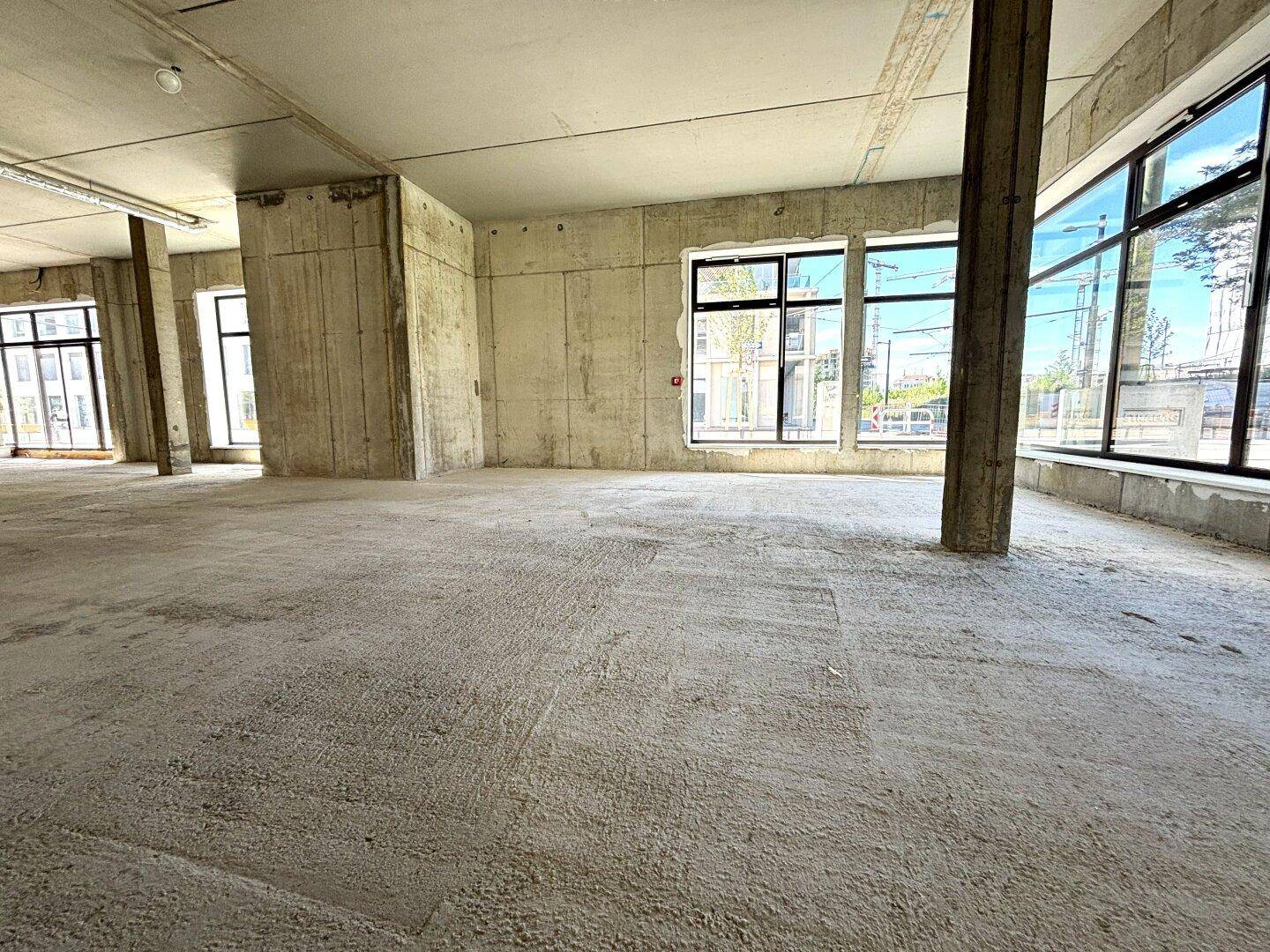 Unfertiger Gewerberaum mit Betonboden, hohen Decken und großen Fenstern, die viel Licht hereinlassen.