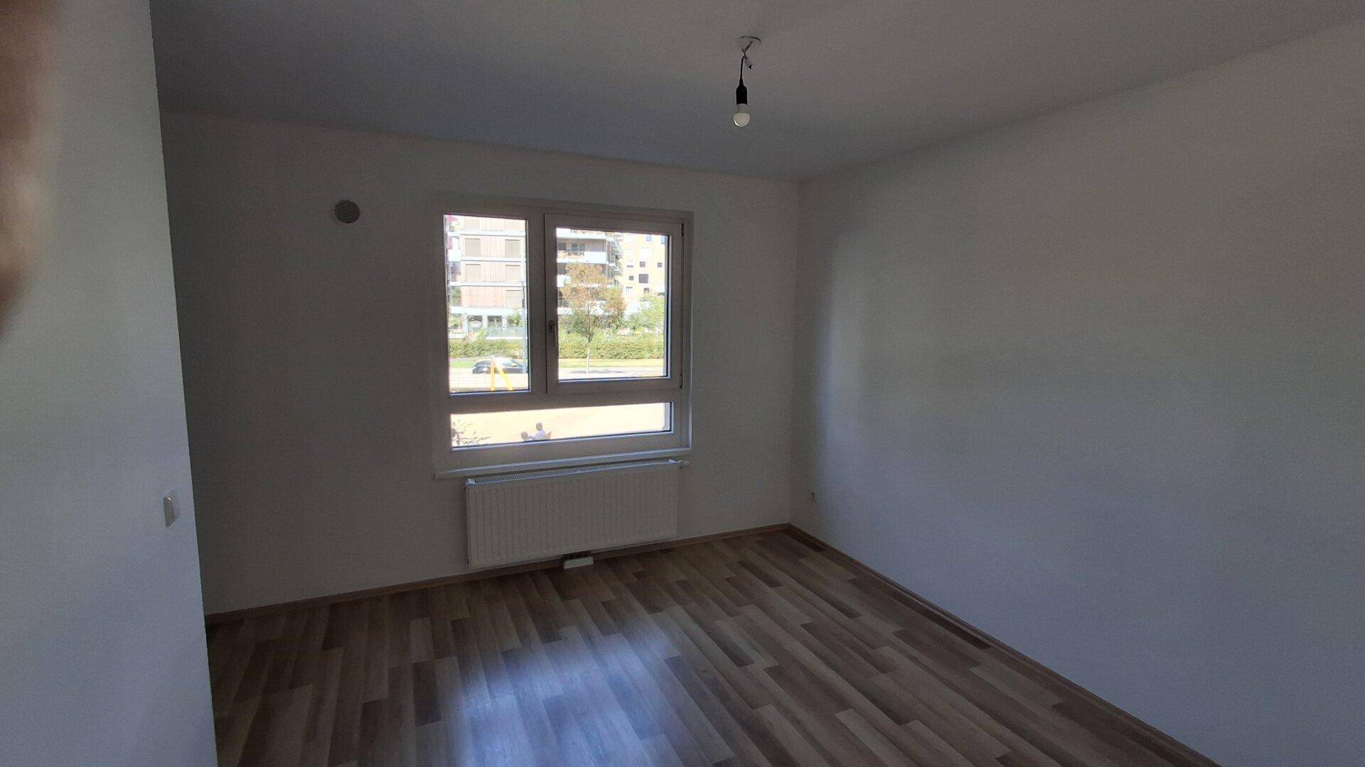 Leeres Zimmer mit Laminatboden, weiß gestrichenen Wänden und einem Fenster mit Blick nach draußen.