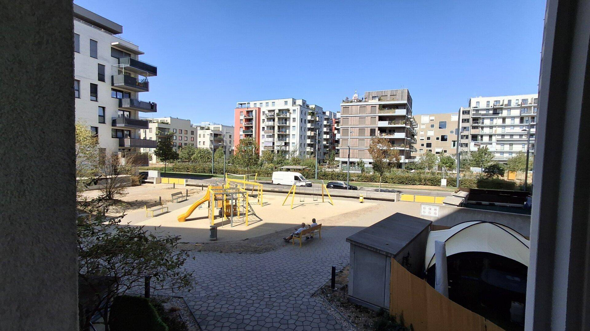 Sonniger Spielplatzbereich mit Rutsche und Schaukeln, umgeben von modernen Wohnkomplexen.