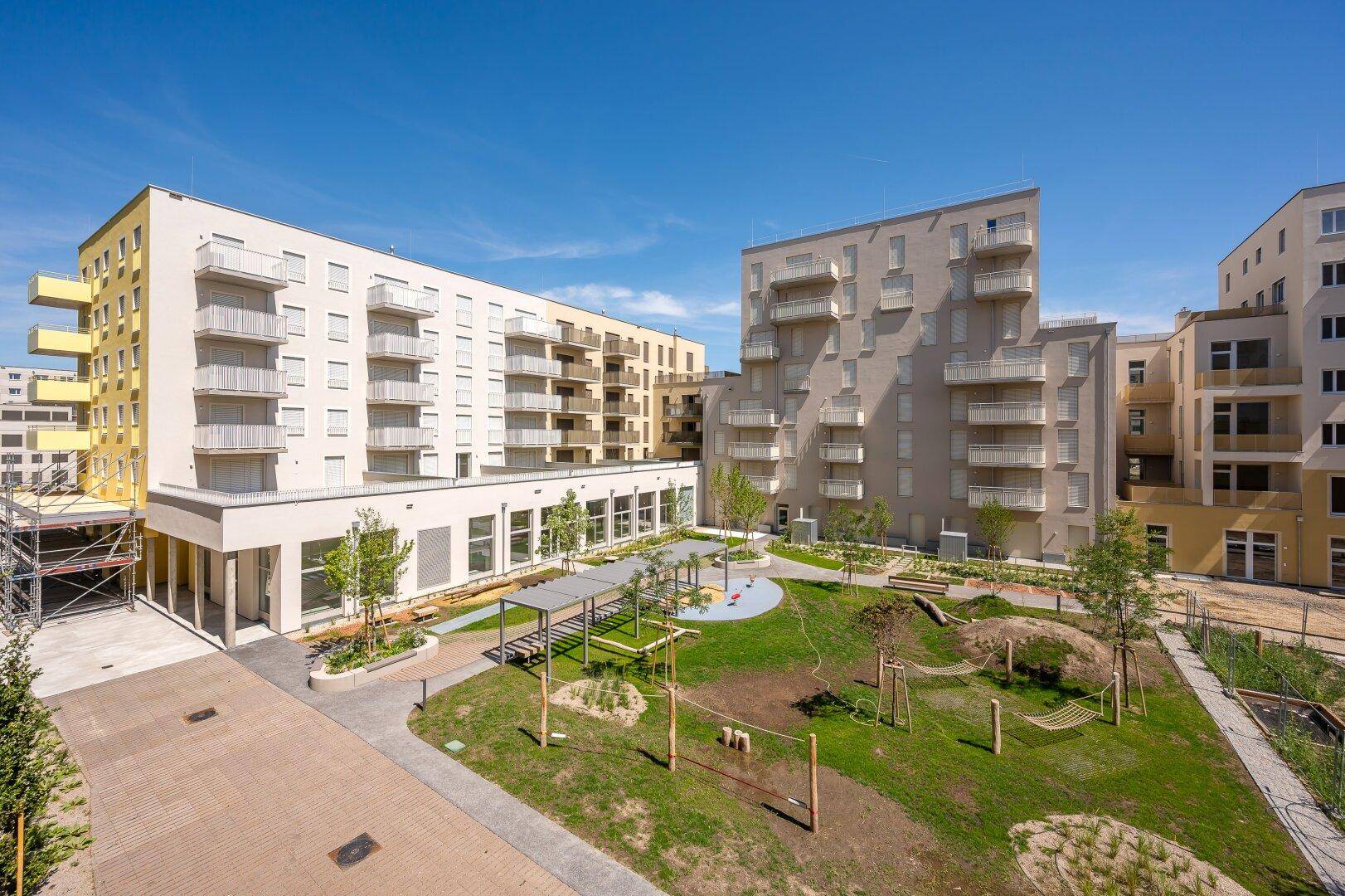 Sonniger Innenhof mit Spielplatz, Hängematte und Grünflächen, umgeben von Neubauten.