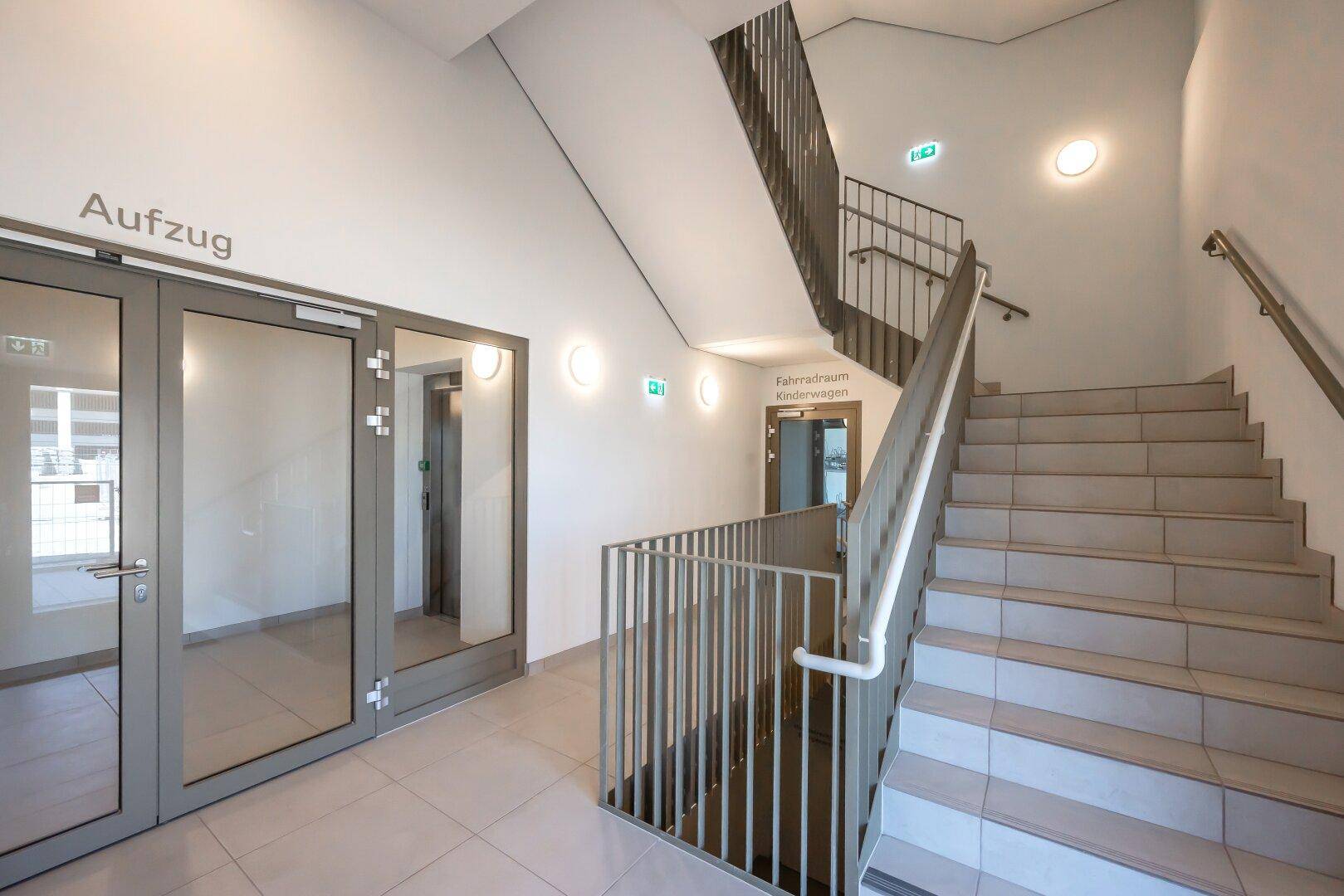Helles Treppenhaus mit Aufzug und Zugang zu den Wohnungen in einem Neubau.