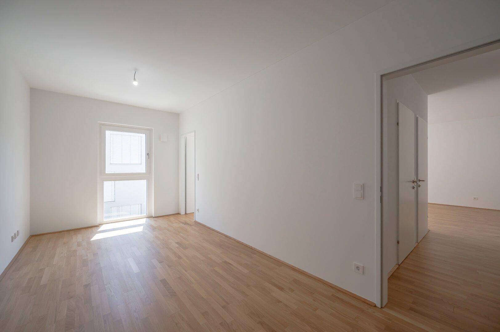 Heller, leerer Raum mit Parkettboden und Fenster, das Zugang zu einem Balkon bietet.