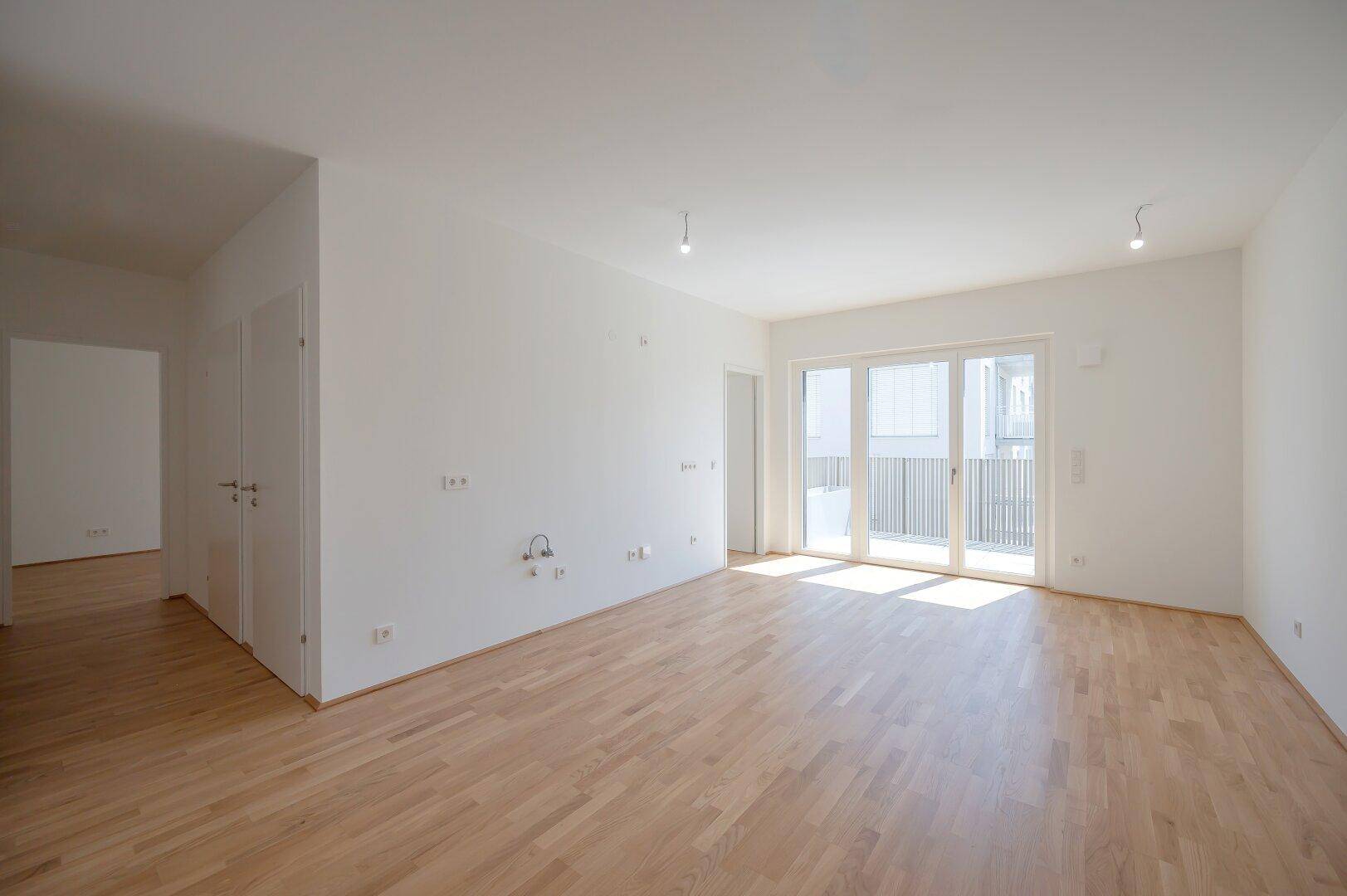 Geräumiges, leeres Wohnzimmer mit hellem Parkettboden und Zugang zu einem Balkon.