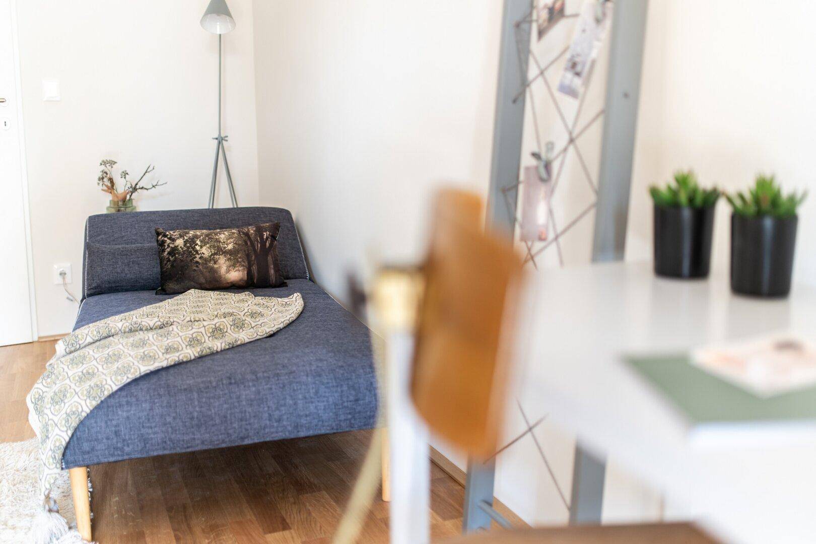 Gemütlicher Bereich mit einer blauen Chaiselongue, Stehlampe und Holzboden, ideal zum Entspannen oder Lesen.
