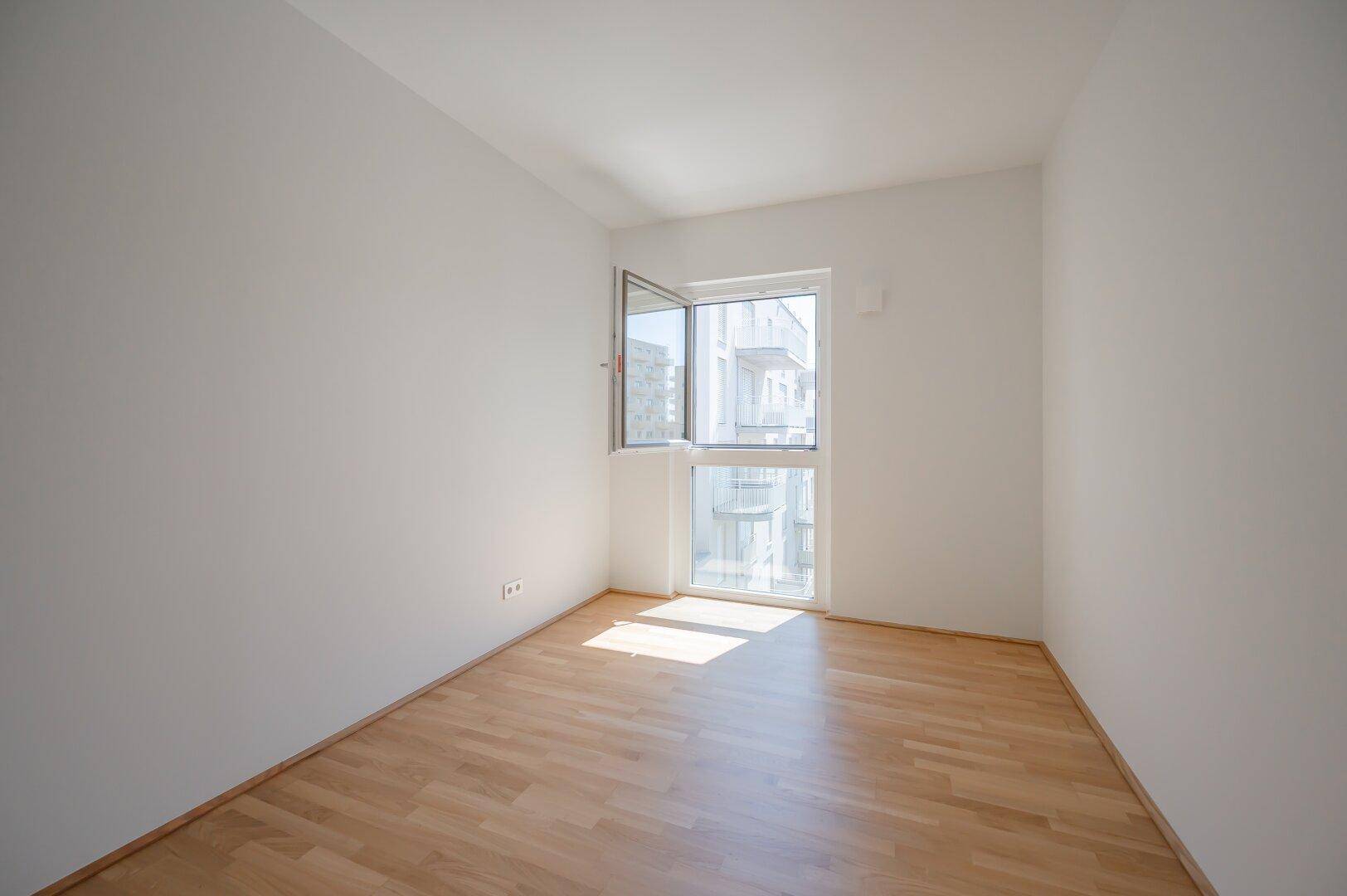 Leeres Schlafzimmer mit hellem Parkettboden und einem großen Fenster für viel Tageslicht.