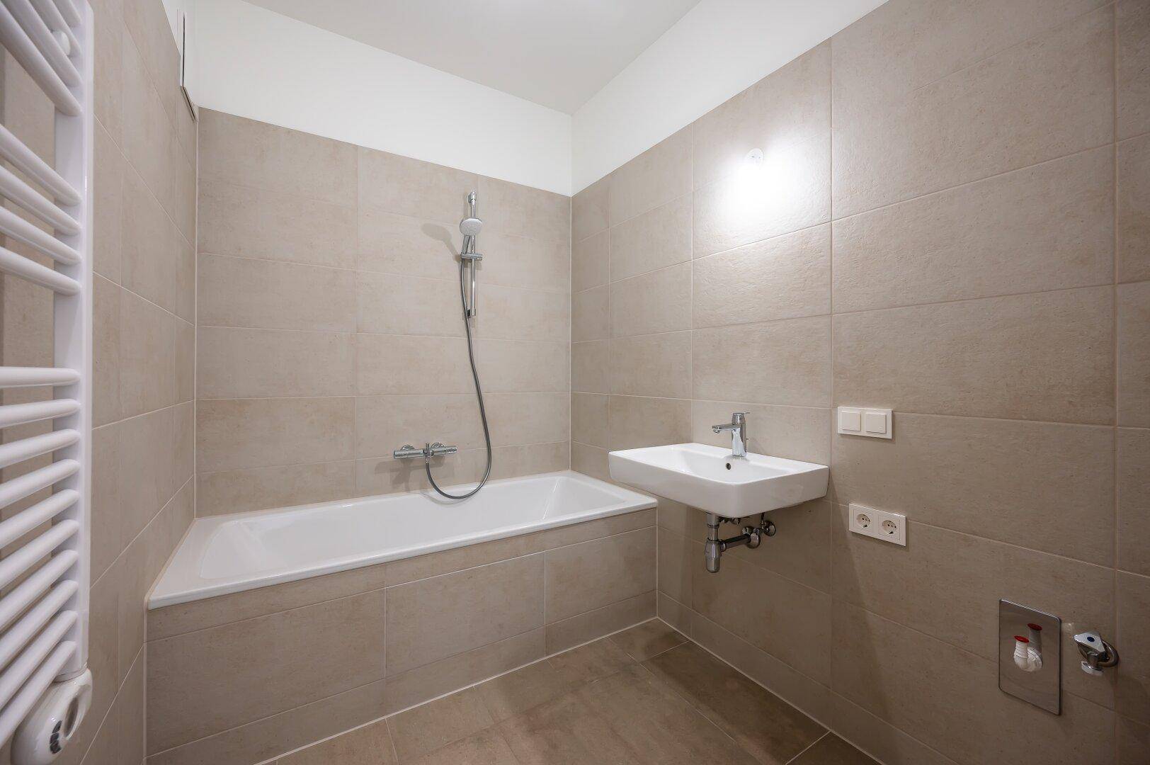 Helles Badezimmer mit Badewanne, Waschbecken und beheiztem Handtuchhalter in beigen Fliesen.