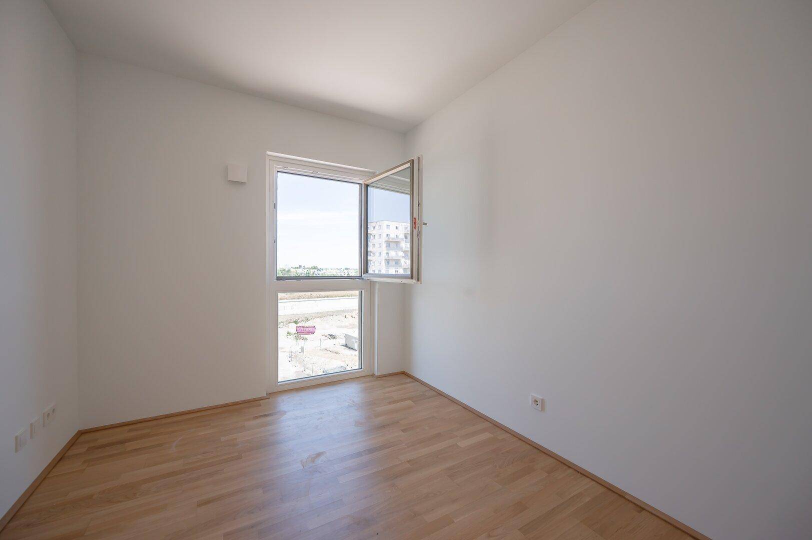 Leeres Zimmer mit Parkettboden und einem großen Fenster, das viel Tageslicht hereinlässt.