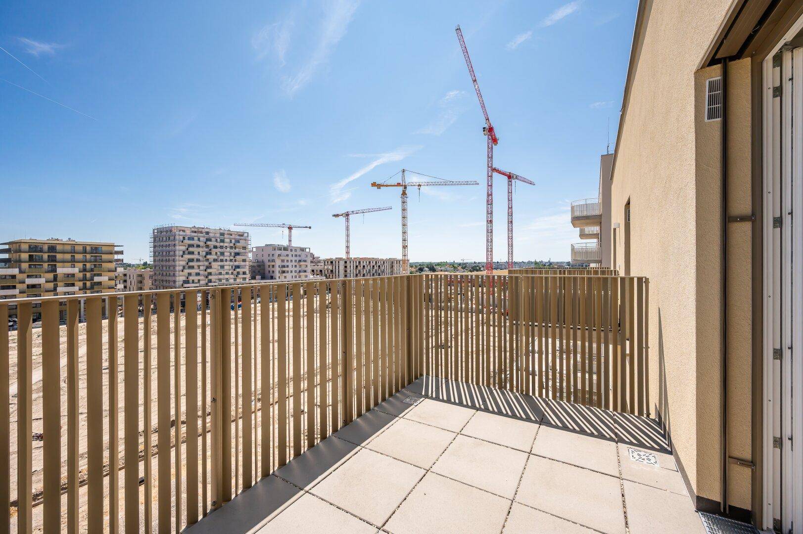 Geräumiger Balkon mit Holzzaun und Blick auf die umliegende Baustelle und Kräne.