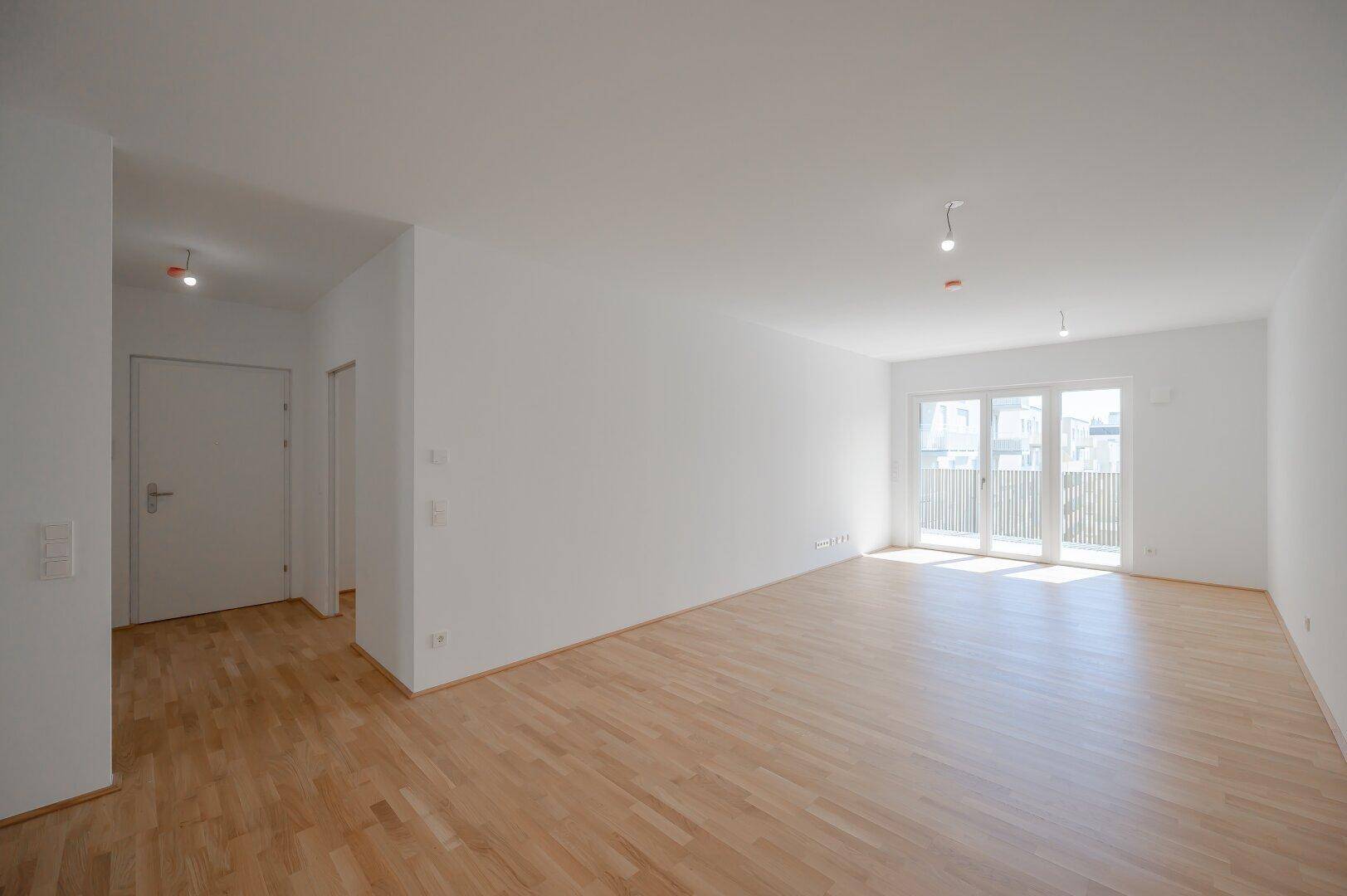 Helles, leeres Wohnzimmer mit Parkettboden und Zugang zu einem Balkon oder einer Terrasse.
